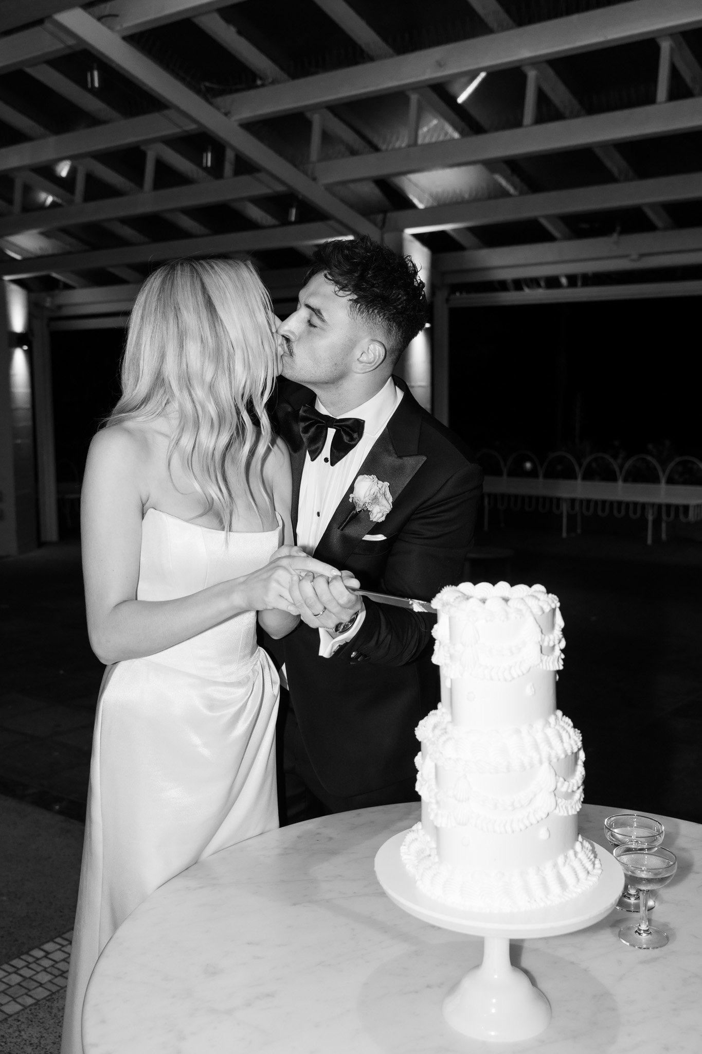 A couple in wedding attire sharing a kiss during their wedding reception, with a wedding cake and glasses on a table in front of them.