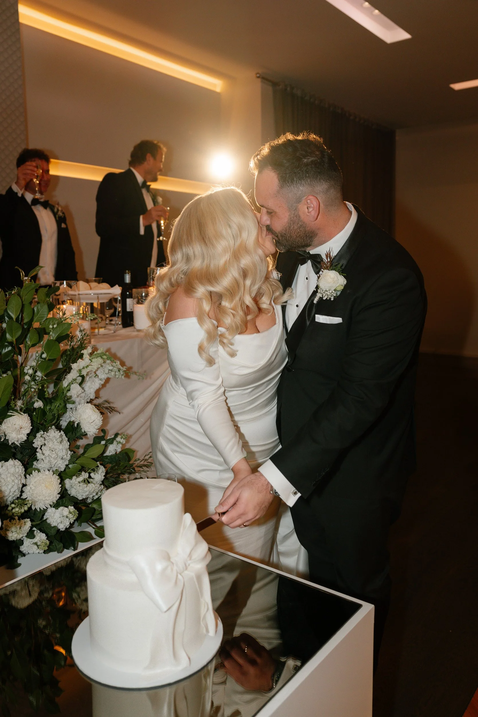 A newlywed couple shares a kiss while cutting their wedding cake at a reception, with friends in tuxedos in the background.
