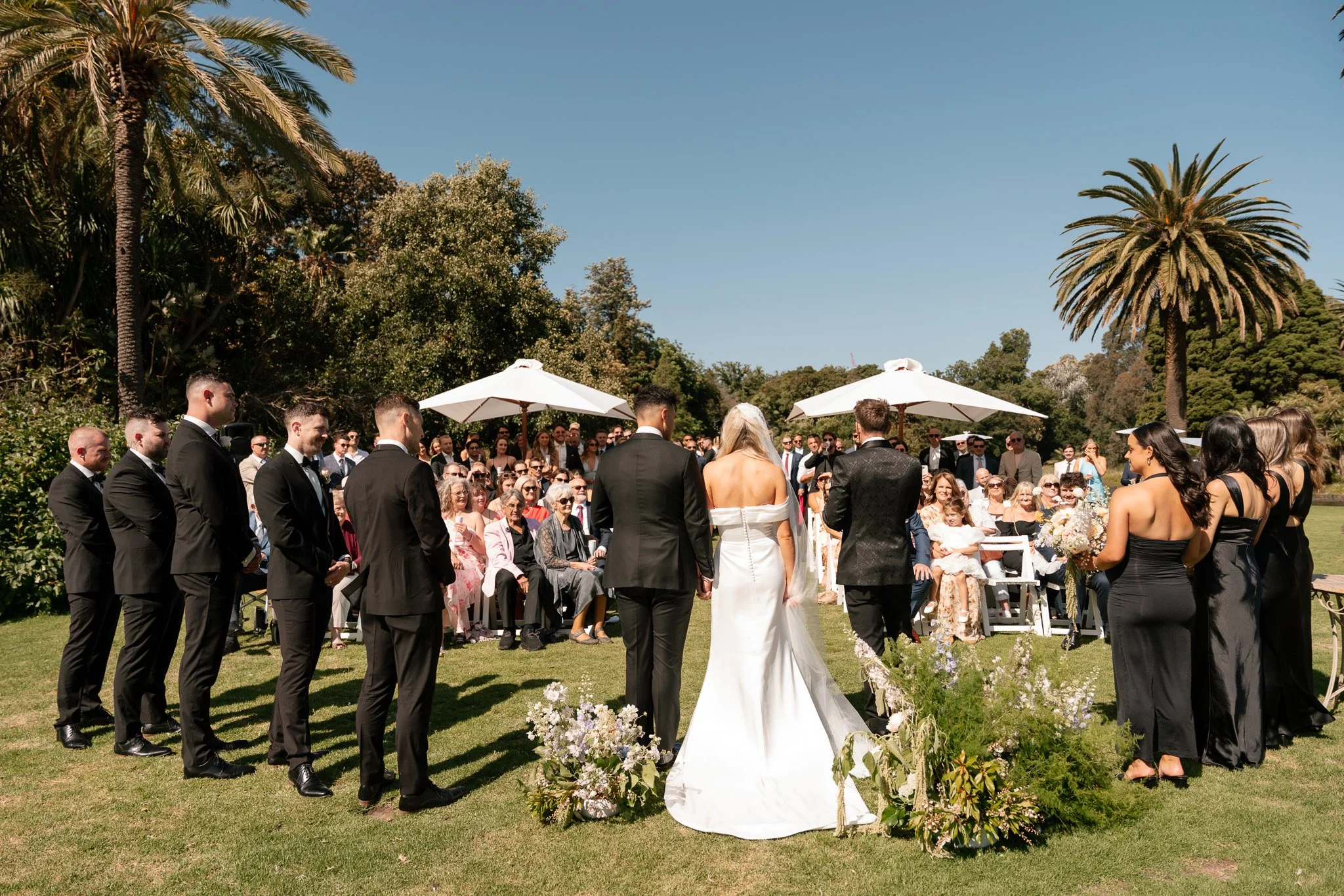 Outdoor wedding ceremony with bride and groom facing guests, surrounded by bridesmaids and groomsmen, under white umbrellas and tall palm trees.