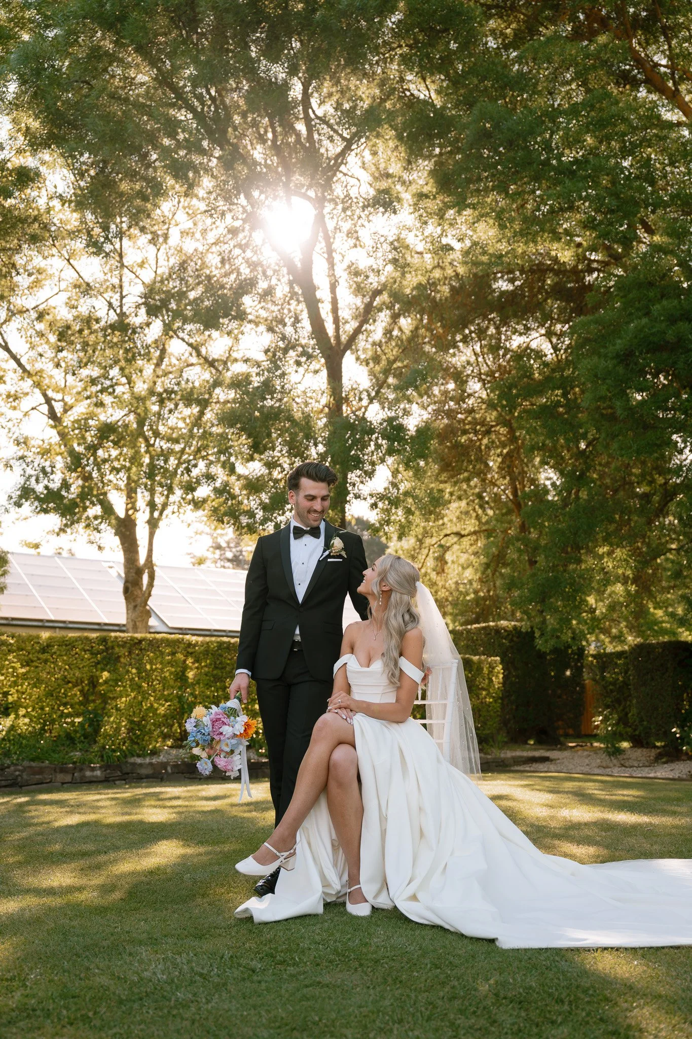 A bride and groom standing outdoors on a grassy yard during sunset, with the bride seated and the groom standing beside her, holding a bouquet of flowers. The bride is wearing a white wedding dress and veil, and the groom is in a black tuxedo with a 