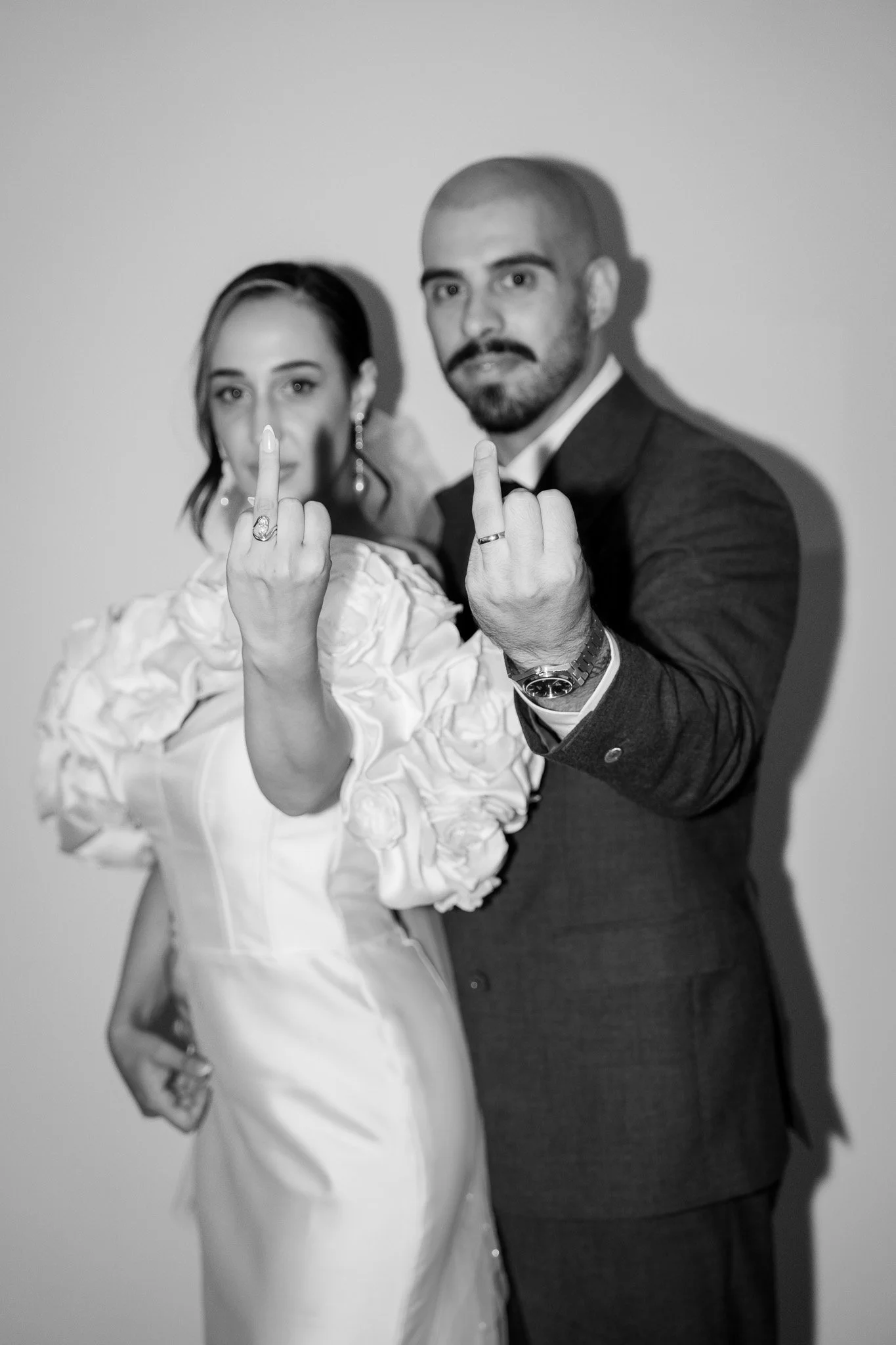 Black and white photo of a woman and man in formal attire, both showing middle fingers to the camera.