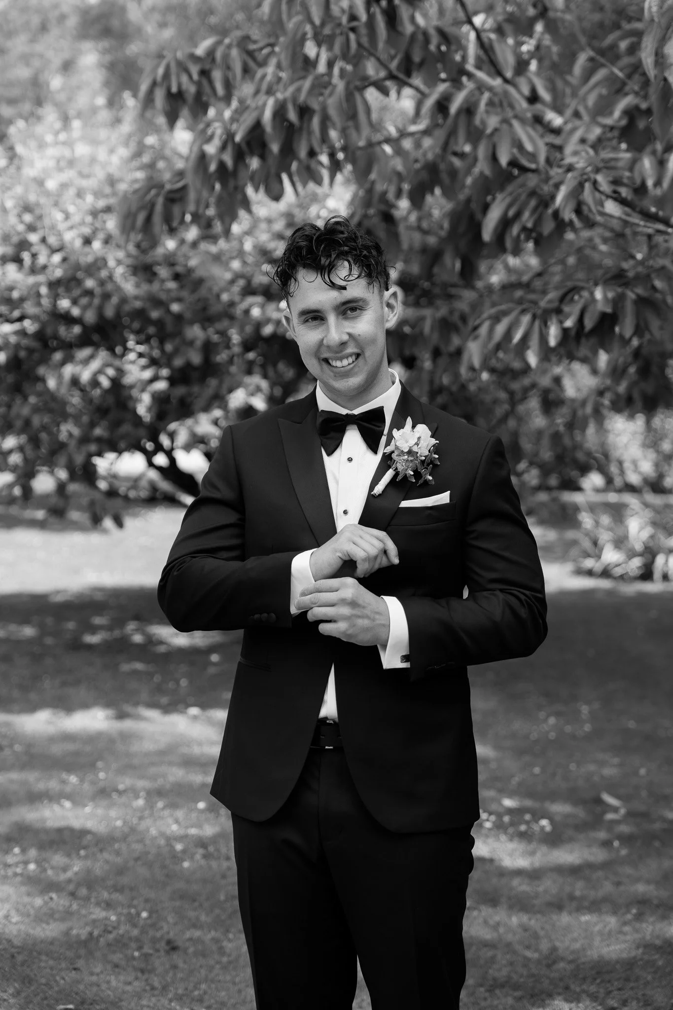 A young man in a tuxedo standing outdoors, smiling, with a boutonniere on his lapel.
