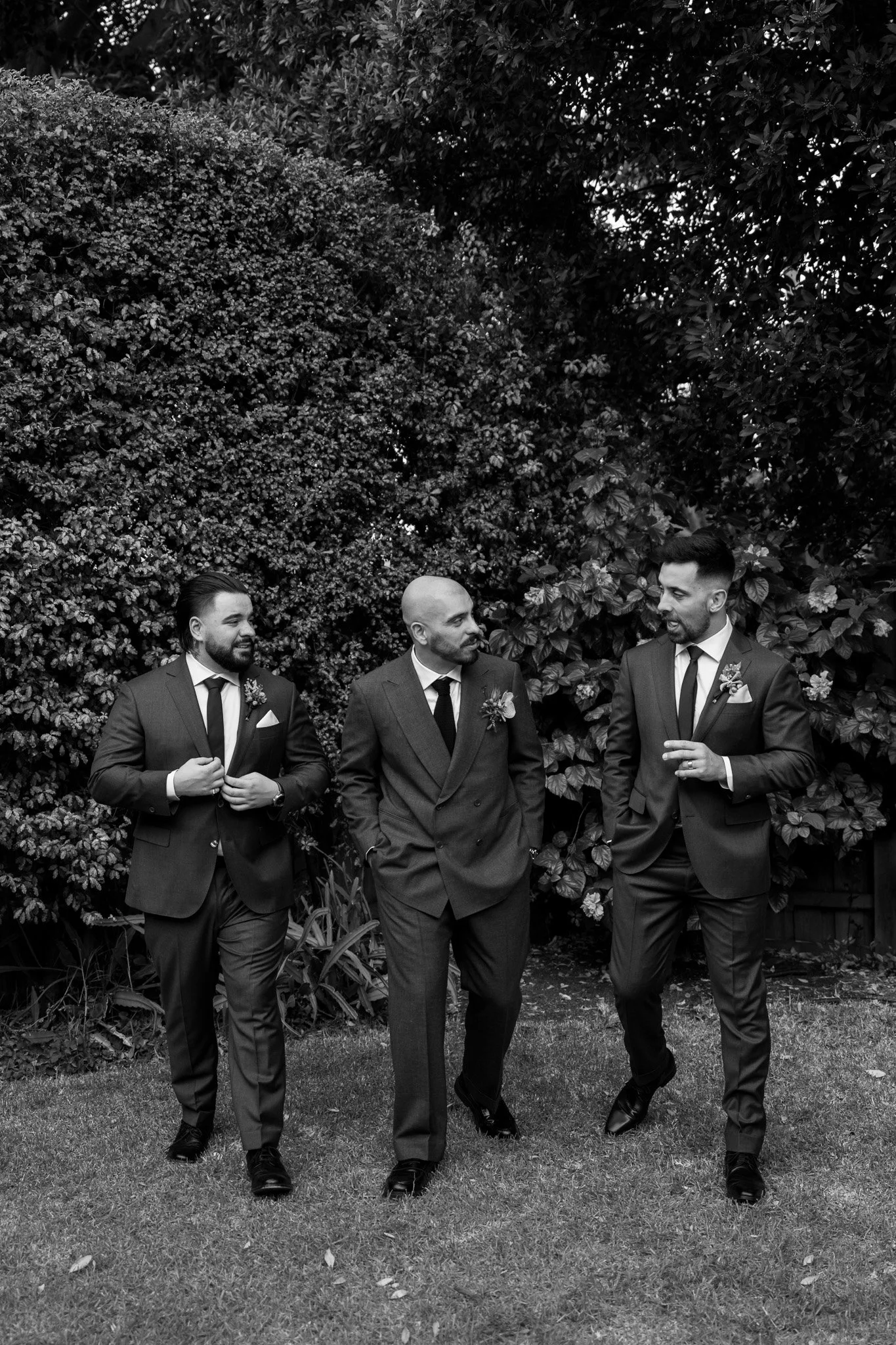 Black and white photo of three men in suits walking and talking outdoors in a garden with bushes and trees.