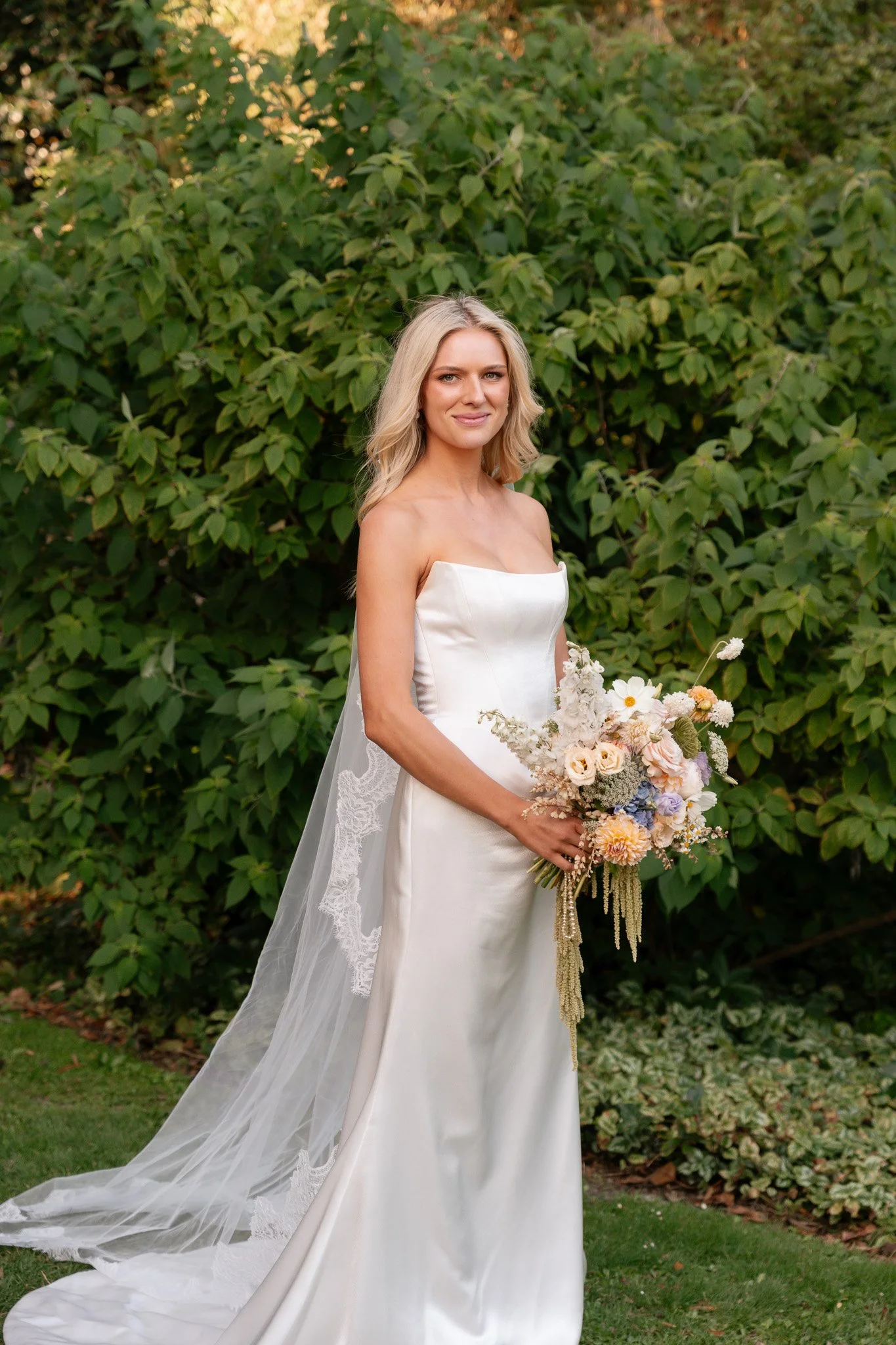 Bride in a white wedding gown holding a bouquet of pastel-colored flowers, standing outdoors in front of green bushes.
