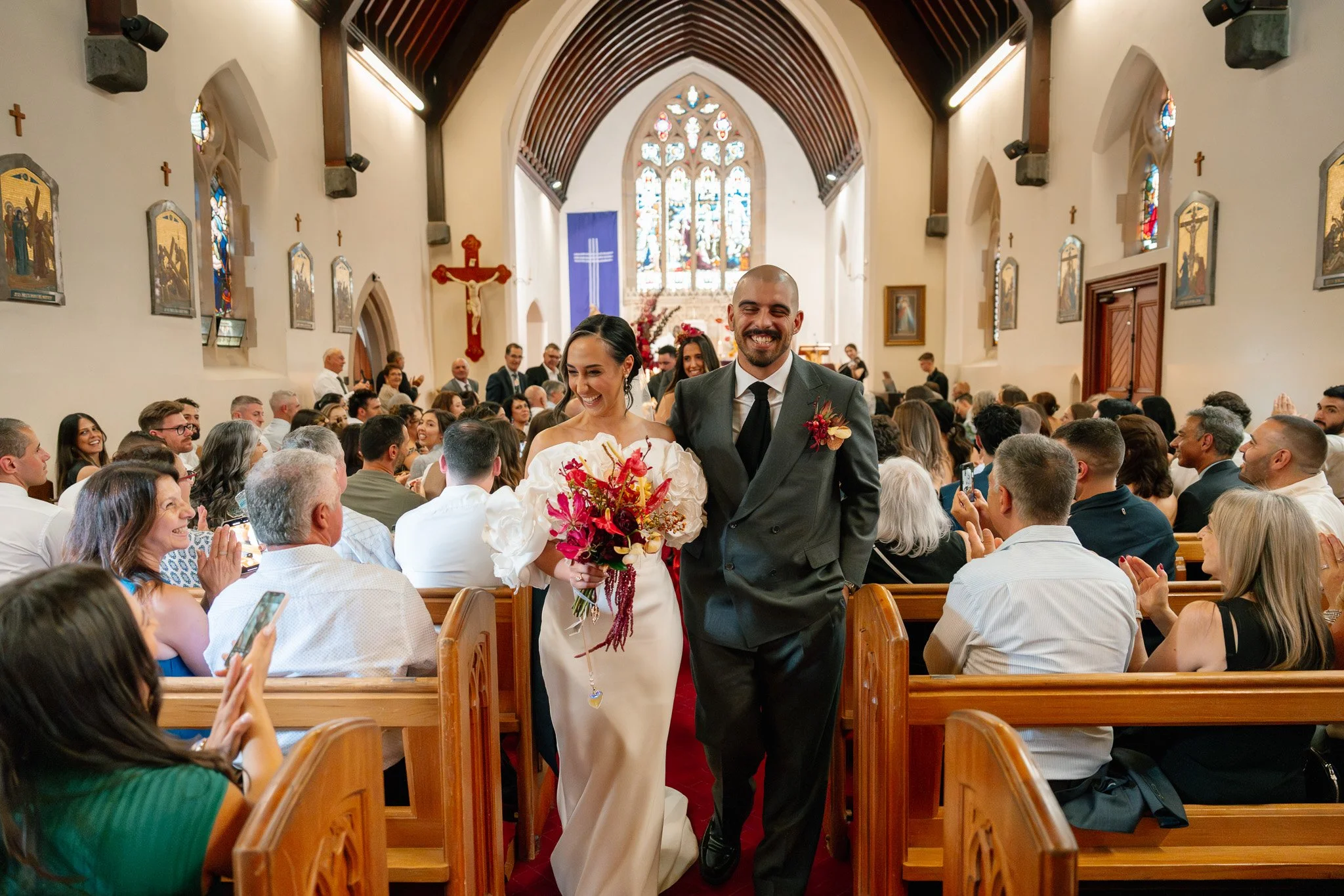Bride and groom walking down the aisle after their wedding ceremony in a church, with guests smiling and taking photos.