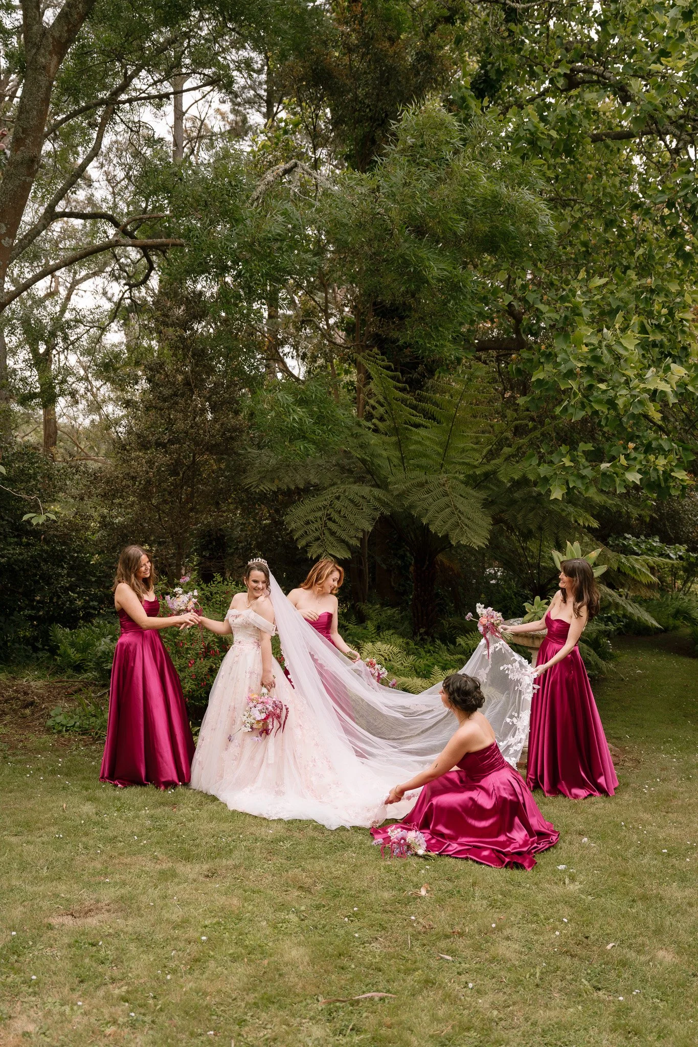 Bride in a wedding dress with veil and tiara, surrounded by five women in matching magenta bridesmaids' dresses, in a lush garden with tall trees and green foliage.