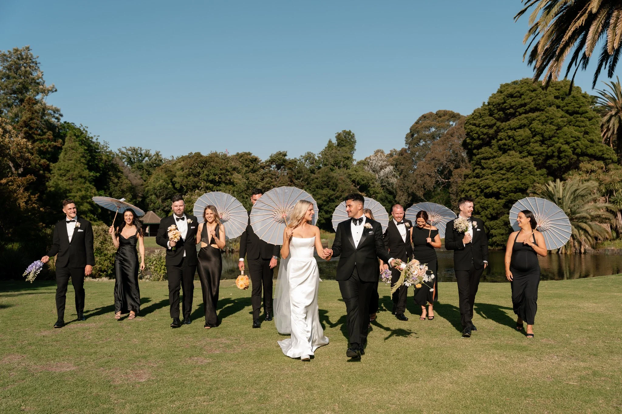 A bride and groom walking hand in hand outdoors with wedding party. Bridesmaids and groomsmen hold umbrellas and flowers on a sunny day surrounded by trees.