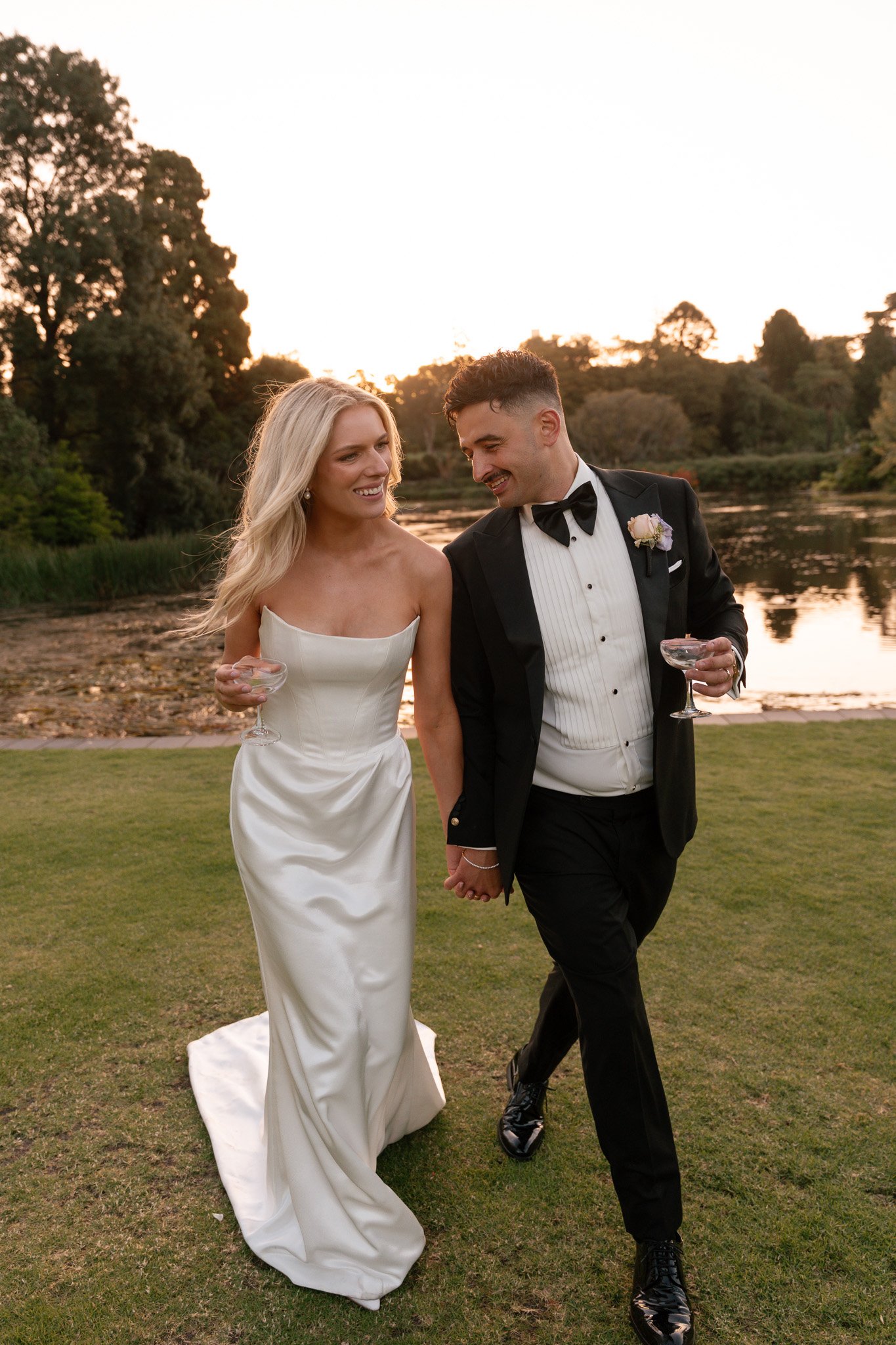 A bride and groom walking hand in hand outdoors at sunset, holding champagne glasses, smiling, in formal wedding attire near a river with trees in the background.