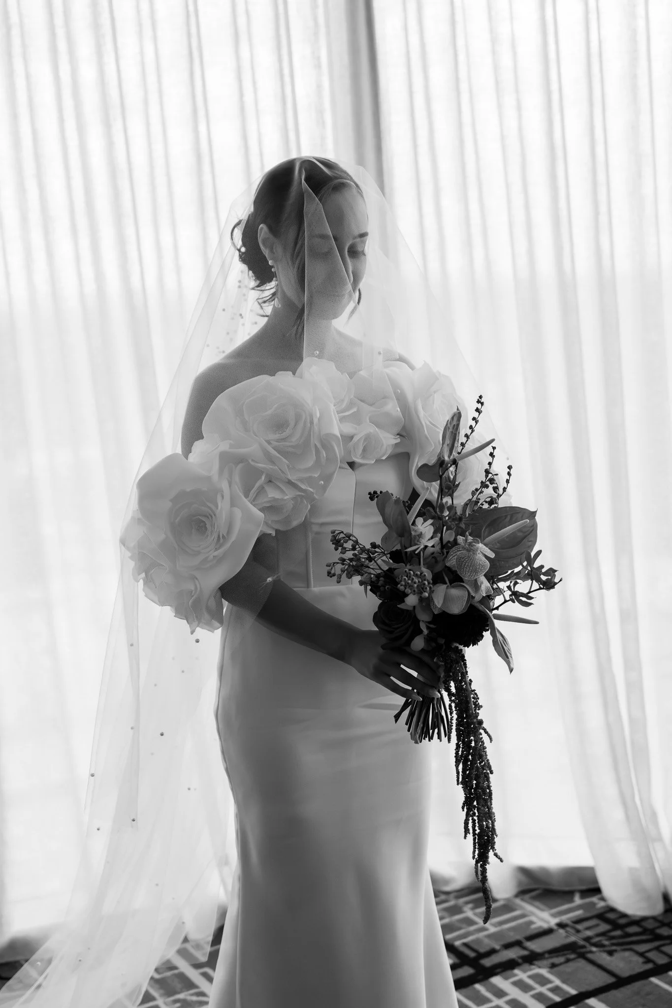Black and white photo of a bride with a veil holding a bouquet of flowers, standing in front of curtains.