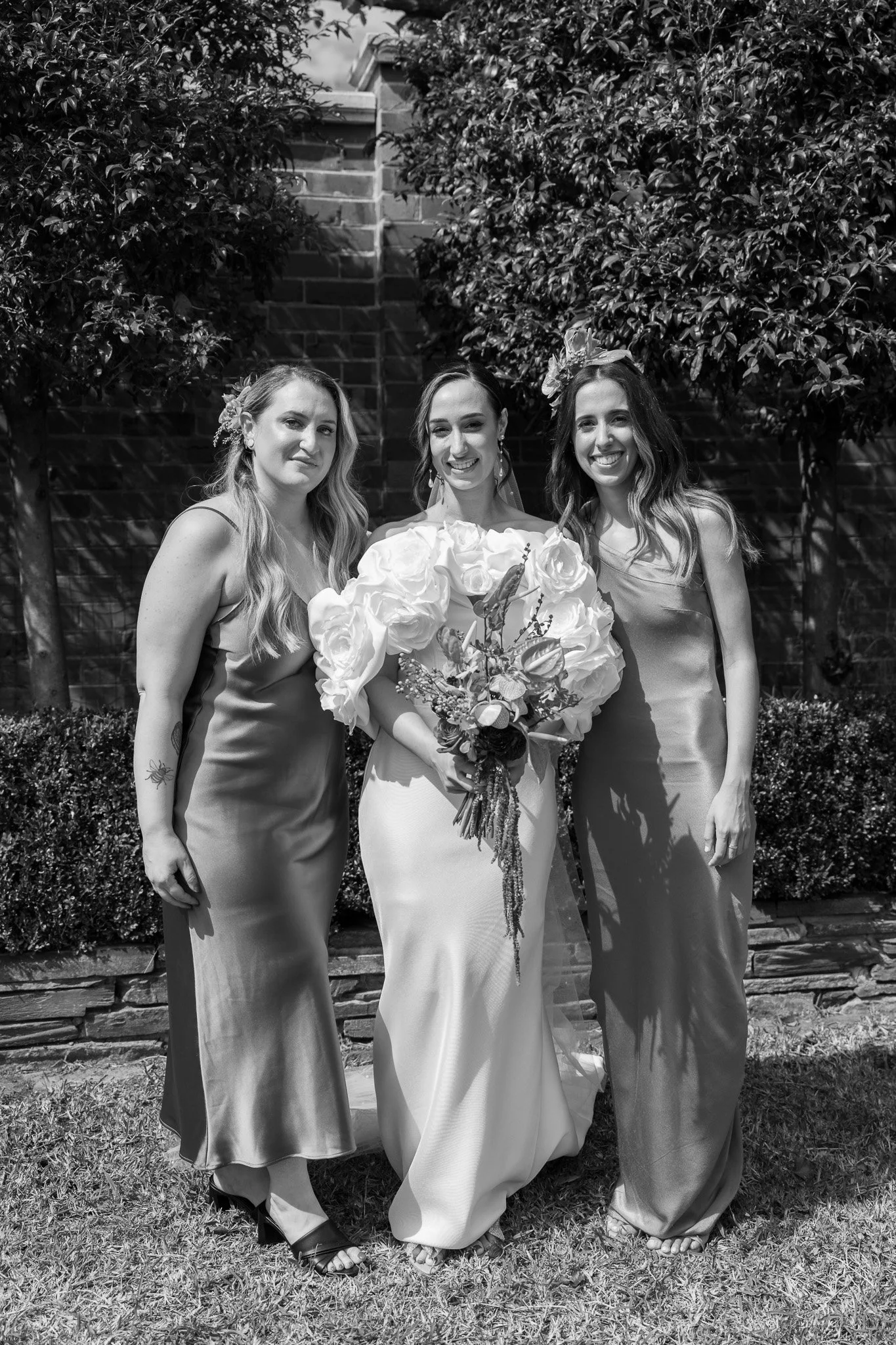 Three women in elegant dresses standing outdoors in front of trees and a brick wall, smiling for the camera, with the woman in the middle holding a bouquet of flowers.