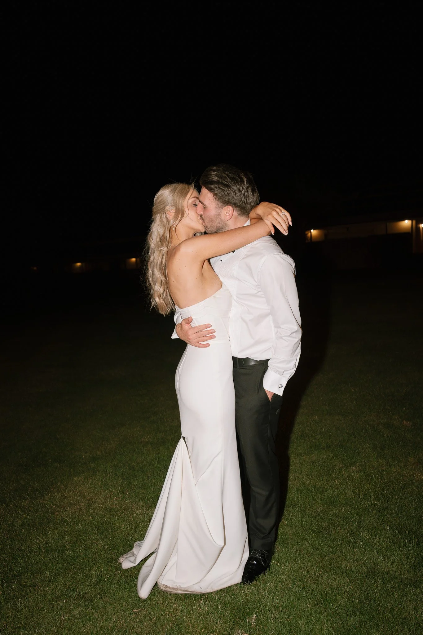 A newlywed couple sharing a kiss at night outside, with the bride in a white strapless gown and the groom in a white shirt and black pants.
