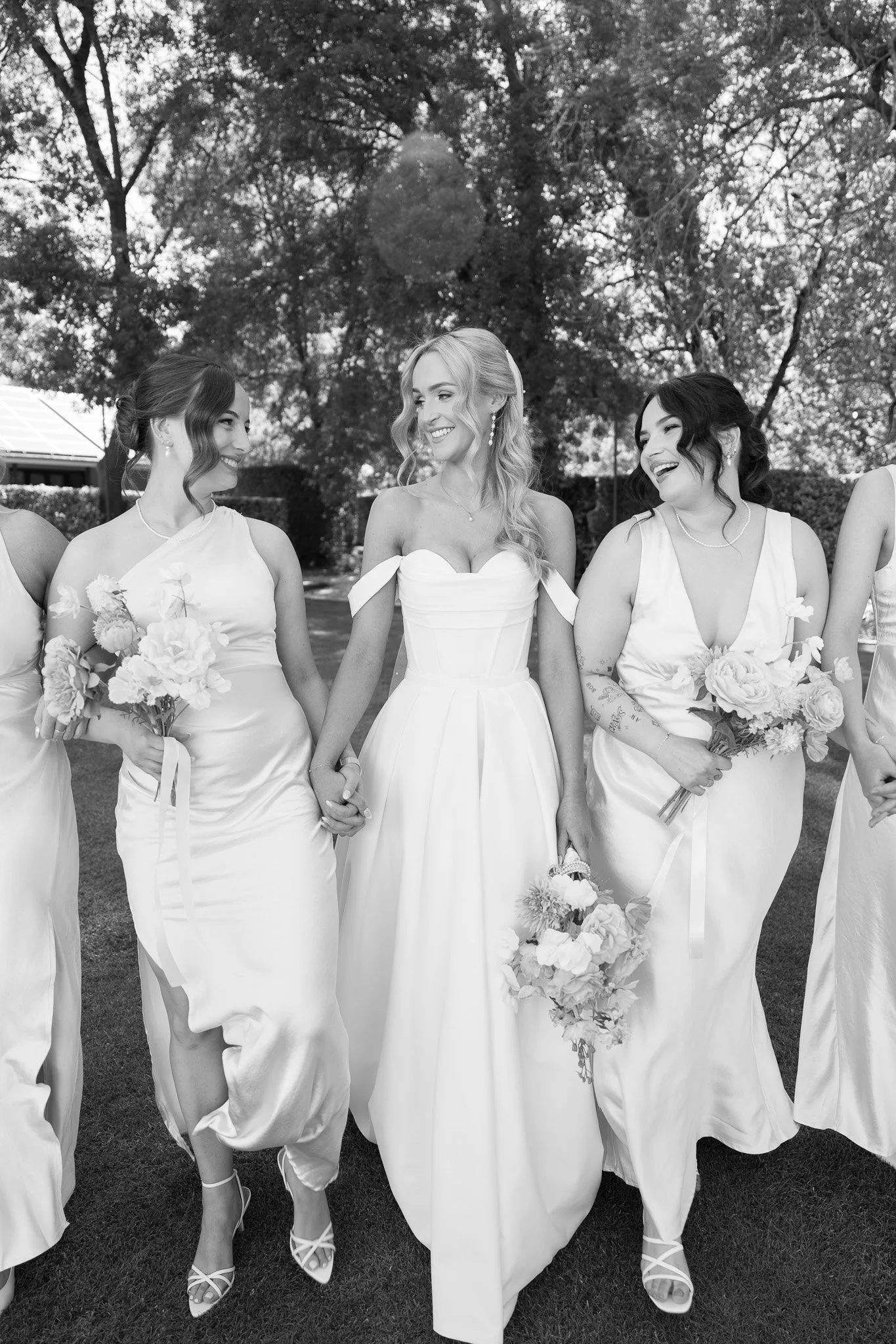 A bride and her bridesmaids holding hands and smiling outdoors on a sunny day, dressed in wedding gowns and holding bouquets.