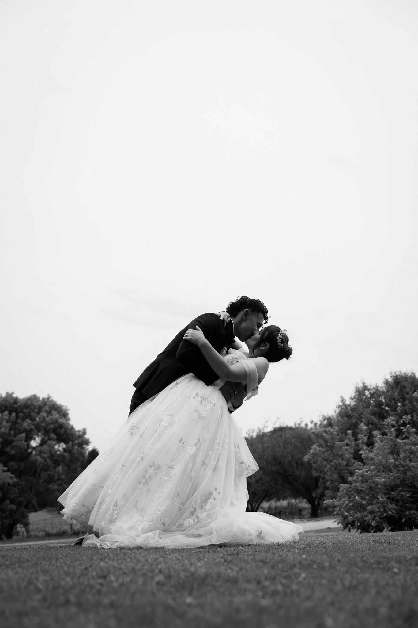 A black-and-white photo of a bride and groom sharing a kiss outdoors, with the groom dipping the bride. The bride is wearing a wedding gown with a long train, and the groom is dressed in a suit. Trees and open sky are in the background.