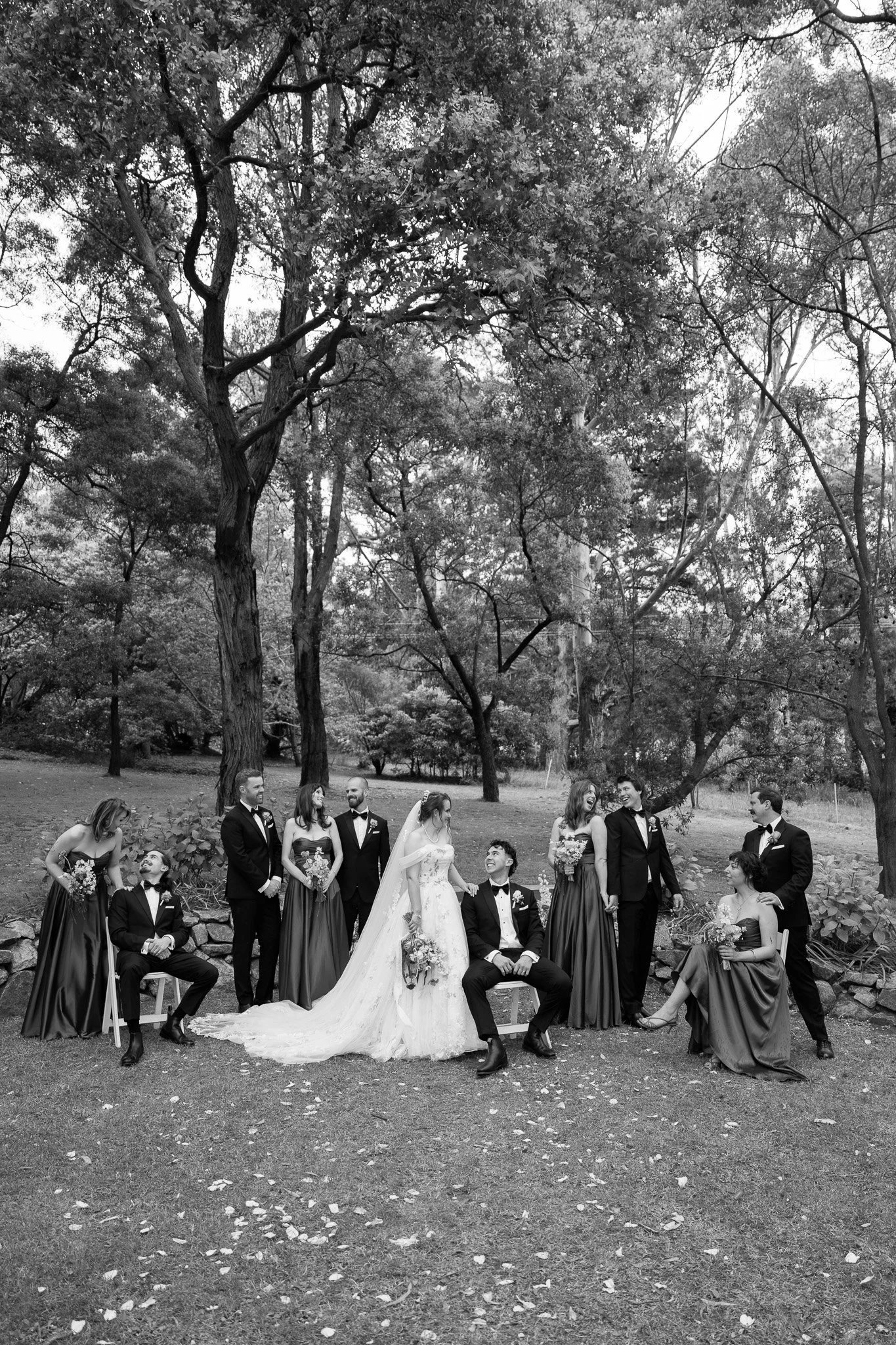 A black-and-white wedding photo of a bride and groom with their wedding party outdoors among trees and grass, with some members sitting and others standing.