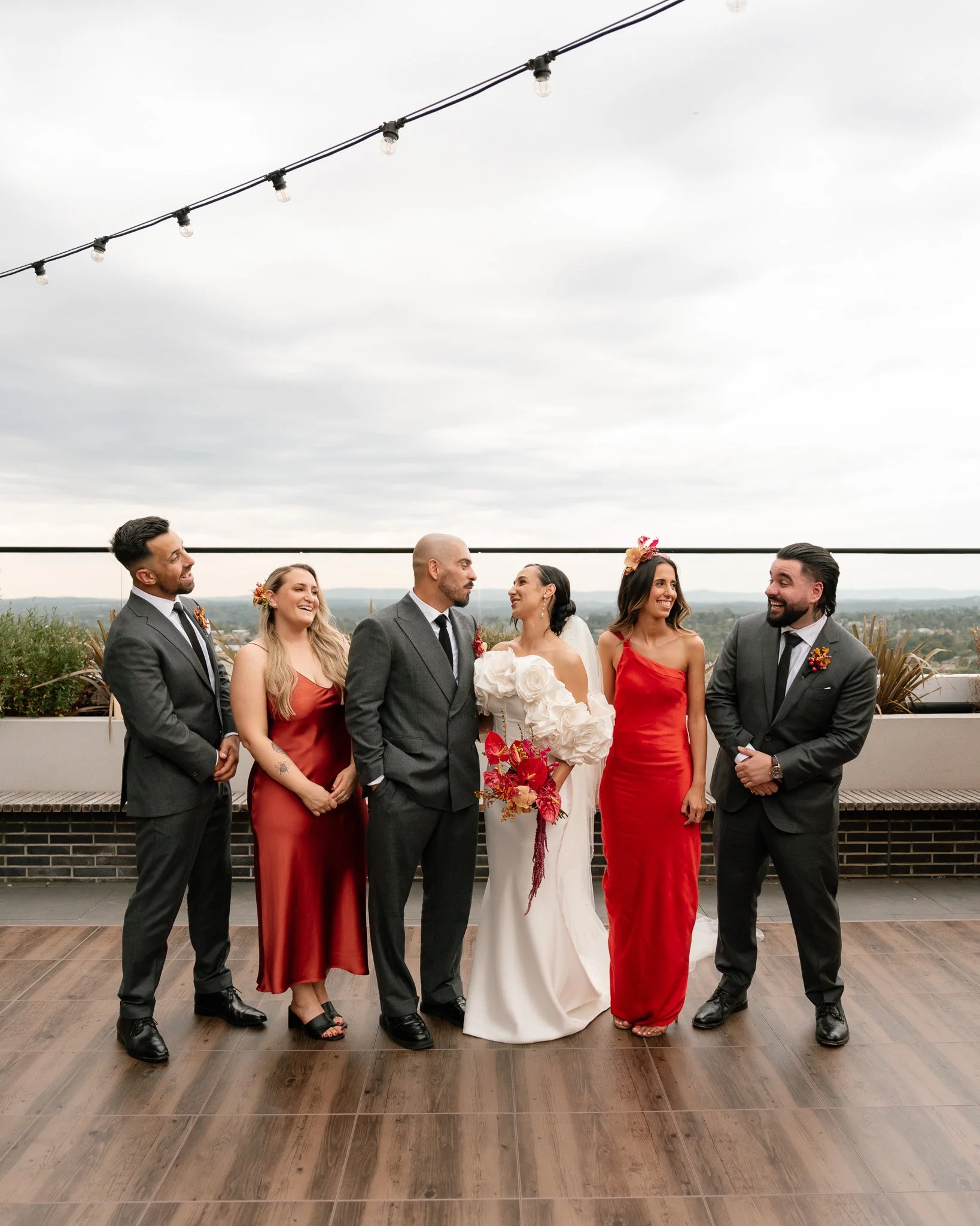 Group of six people, including bride and groom, dressed in formal attire, standing on a rooftop terrace with string lights and a city view in the background, smiling and enjoying the moment.