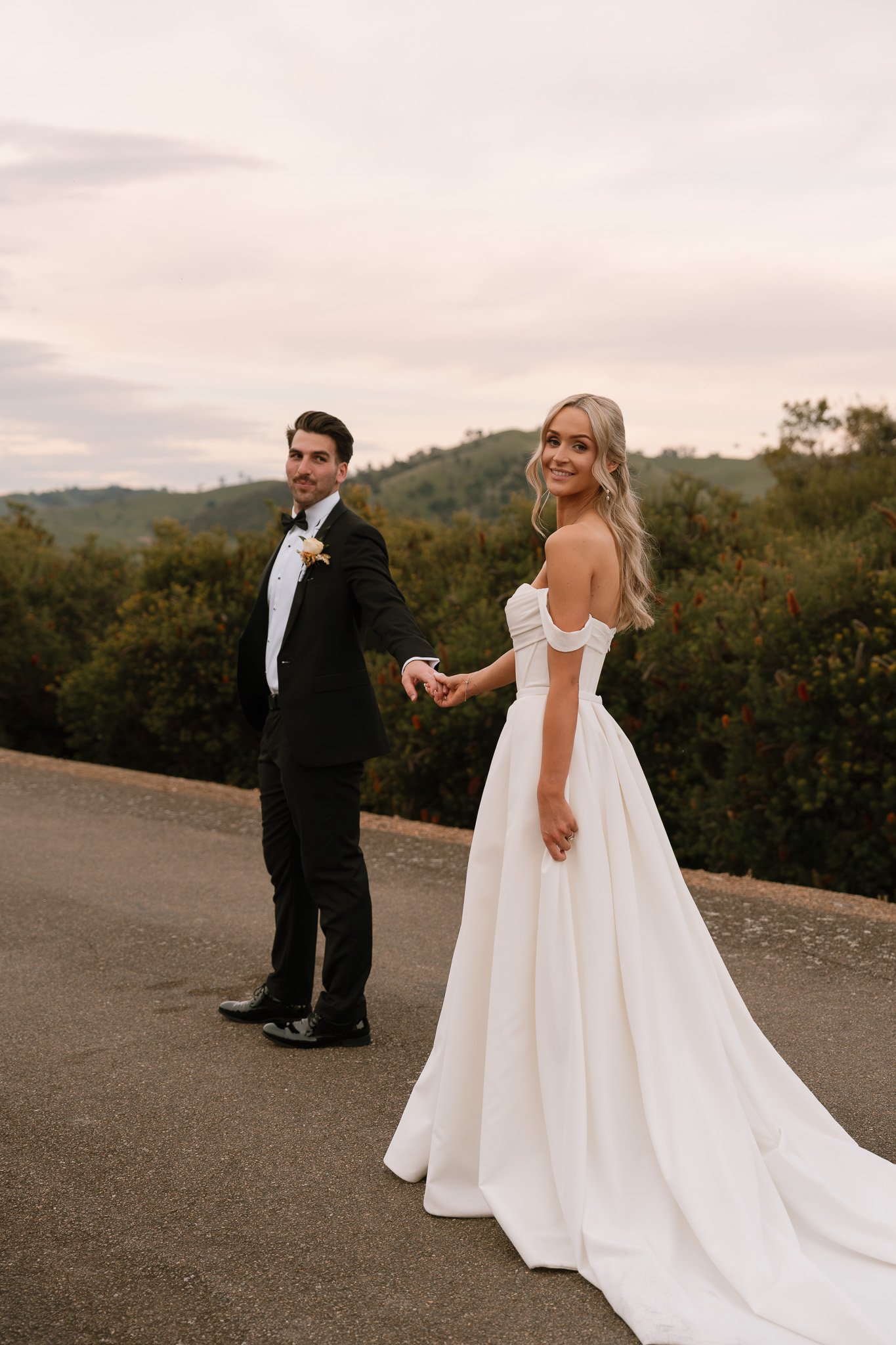 A bride and groom holding hands outdoors on a paved road, with green hills and trees in the background. The bride is in a white off-the-shoulder wedding gown, and the groom is in a black tuxedo.