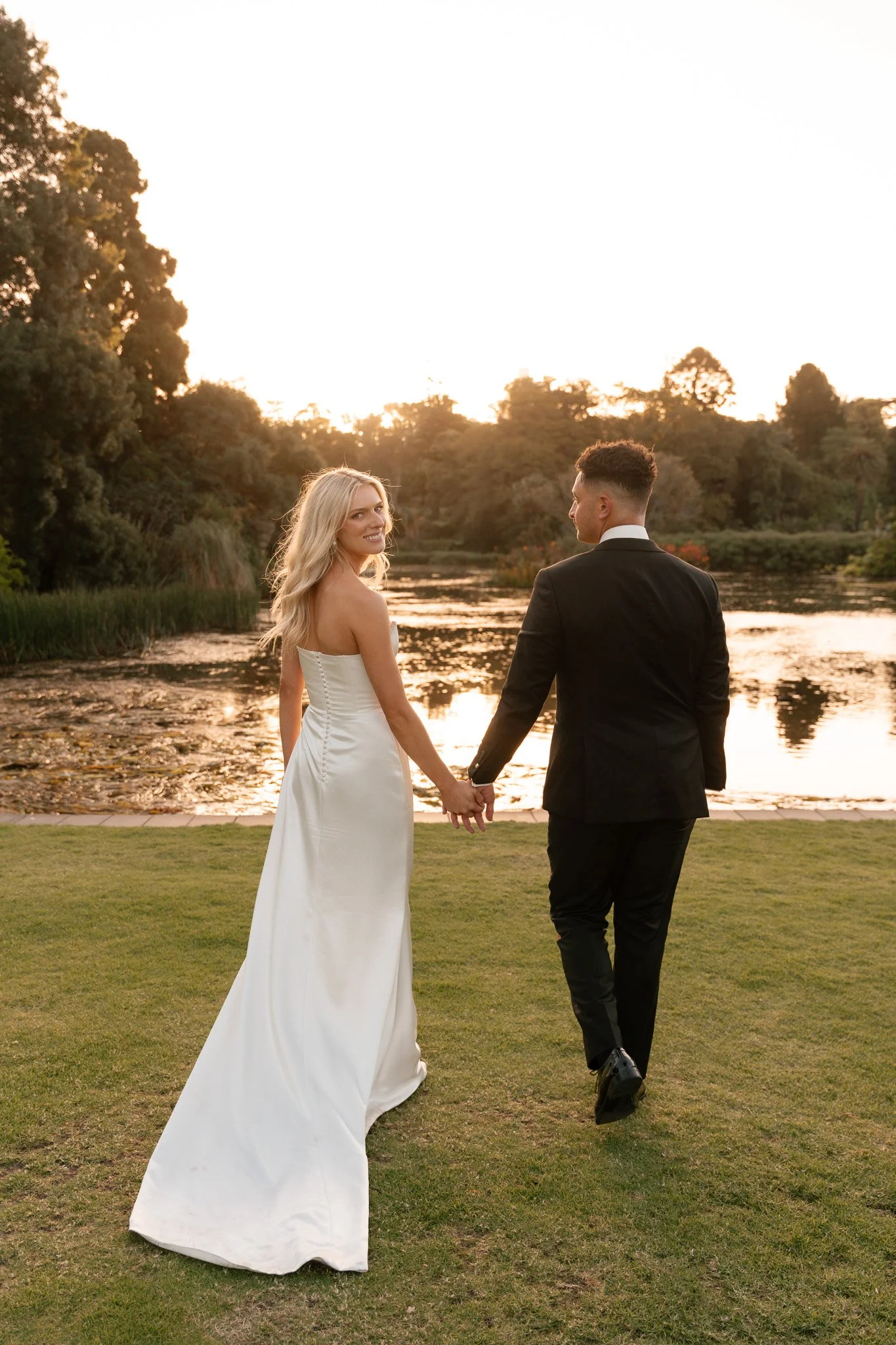 A newlywed couple holding hands and walking on a grassy area near a body of water at sunset, with trees and a bright sky in the background.