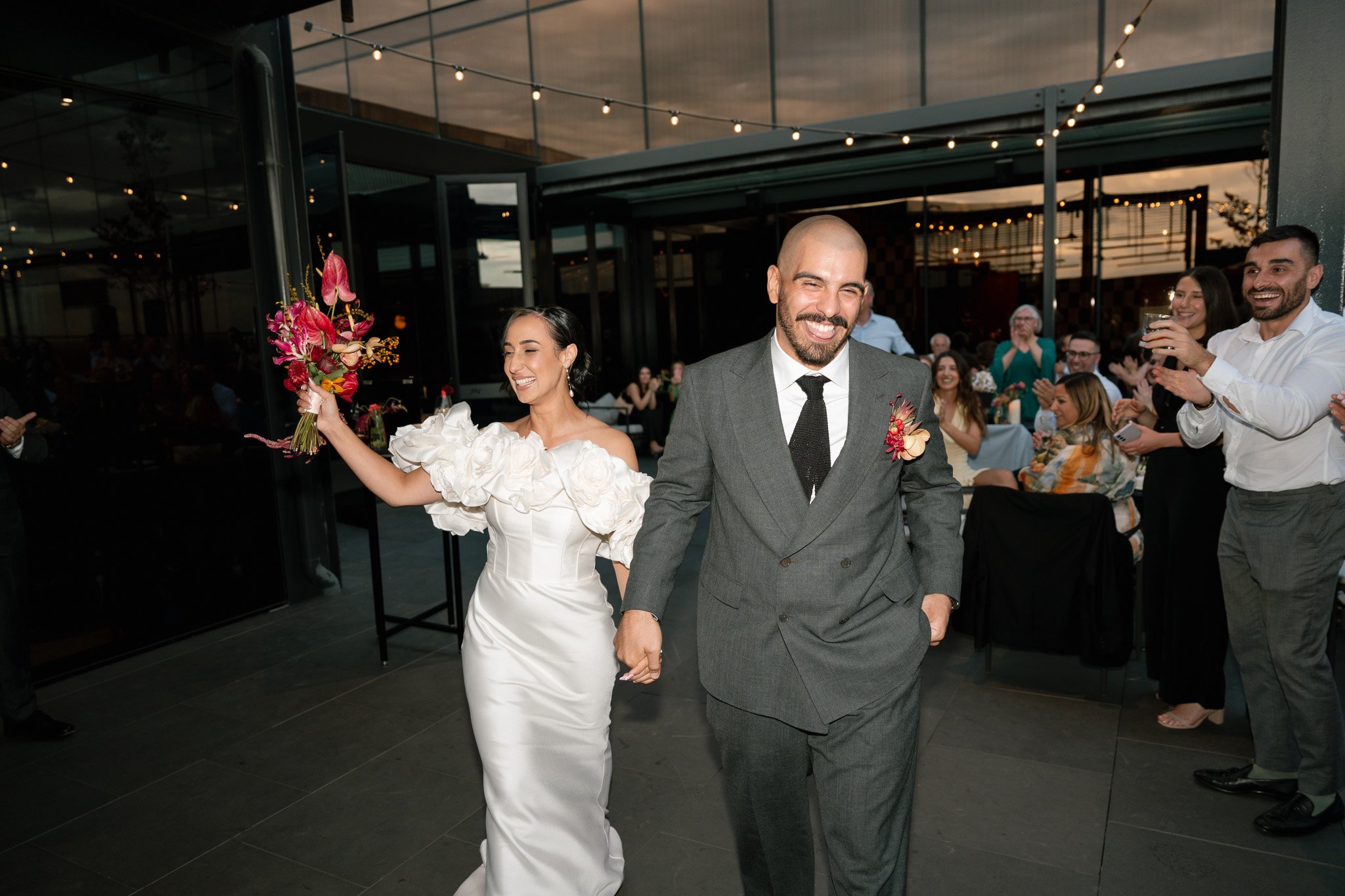 A newlywed couple is walking into their celebration, holding hands, with the bride holding a bouquet of floral arrangements. The bride is wearing a white wedding dress with ruffled sleeves, and the groom is in a gray suit with a black tie and a flora
