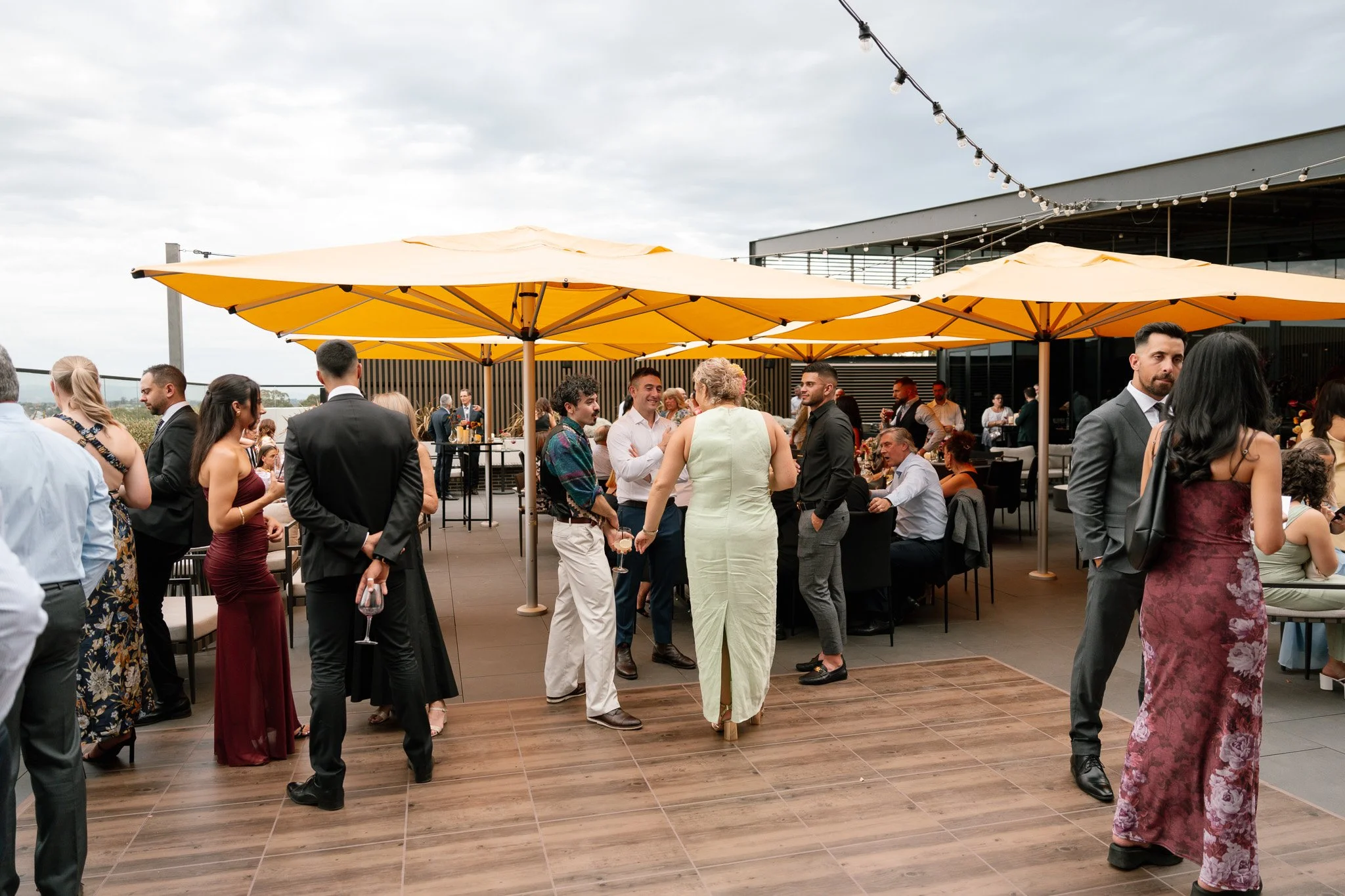 A group of people at an outdoor party or event, socializing and talking under large yellow umbrellas on a rooftop or terrace with some seated guests in the background.