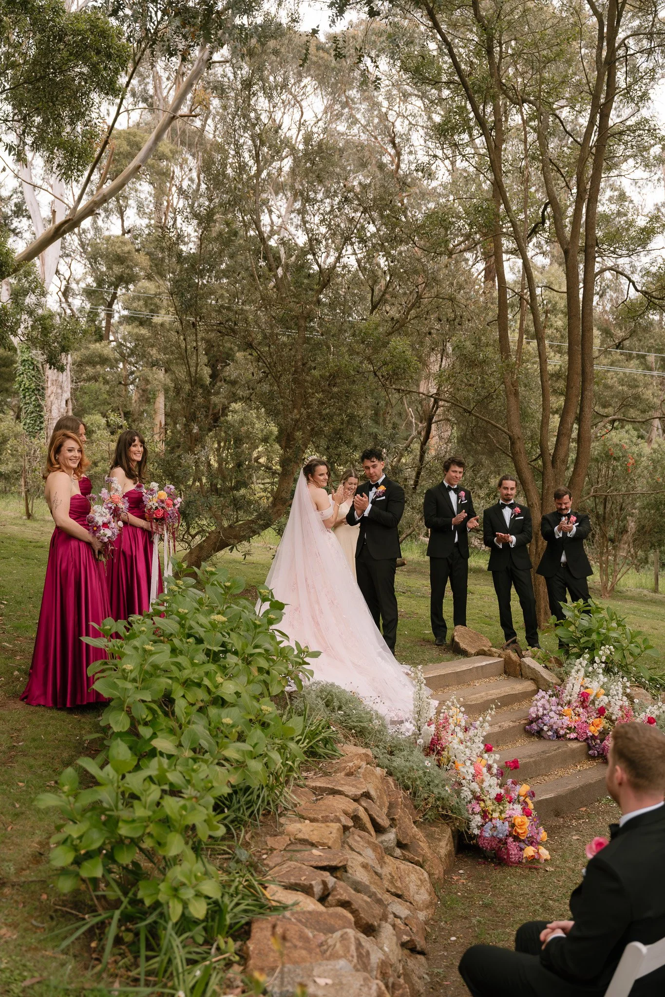 Wedding ceremony outdoors in a wooded area with bridesmaids in magenta dresses, a bride in a white gown, and groomsmen in black tuxedos, floral decorations on steps.