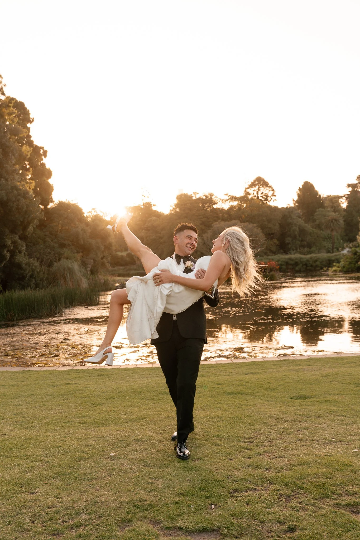 A groom in a tuxedo lifts a bride in a wedding dress and heels, smiling and looking at each other, outdoors near a pond at sunset.