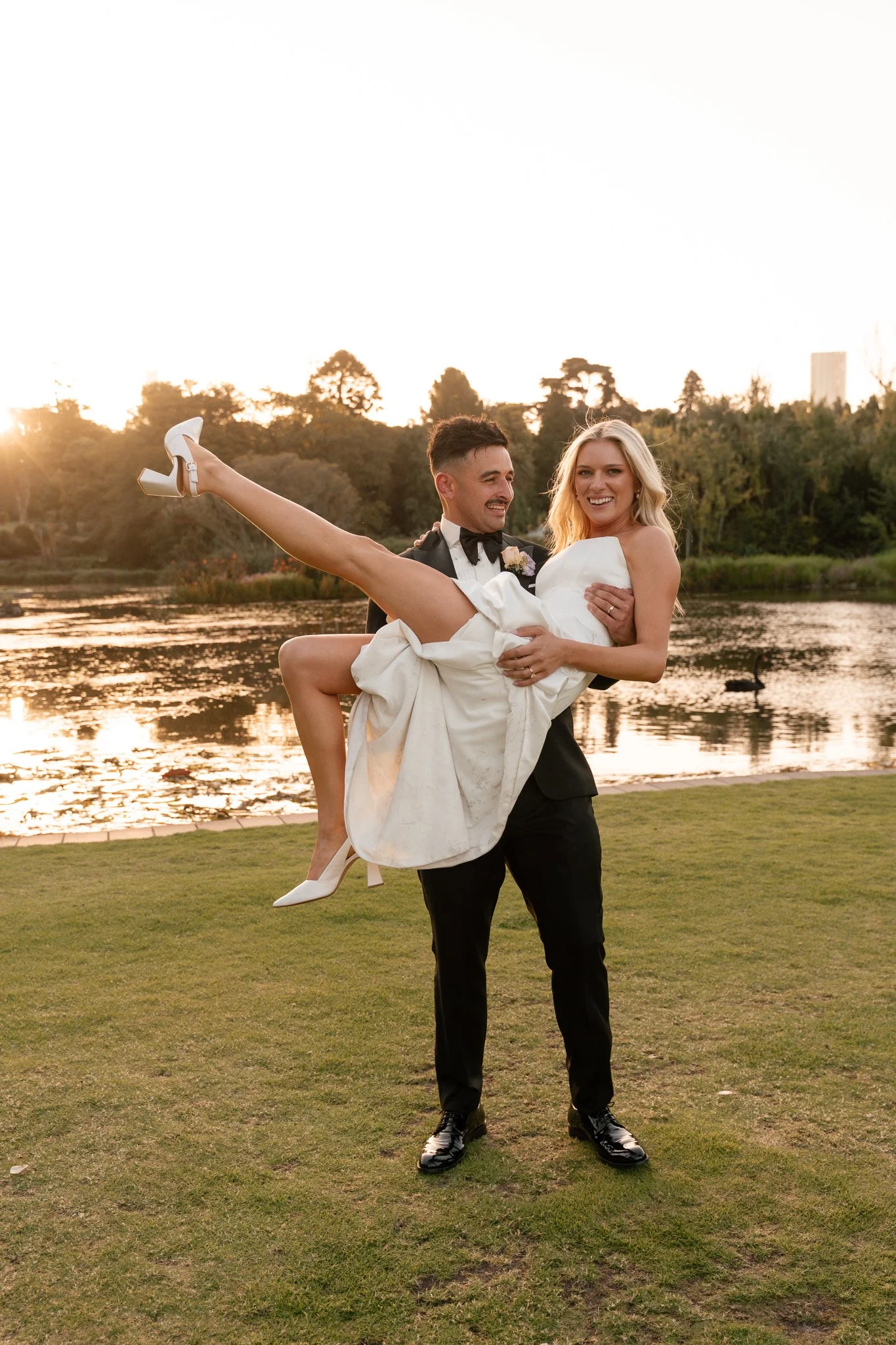 A man in a tuxedo holding a woman in a wedding dress by a lake during sunset.