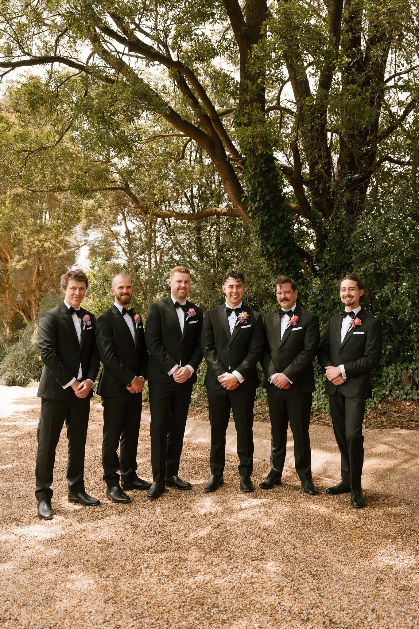Six men in black tuxedos standing outdoors on a gravel path surrounded by trees and greenery.