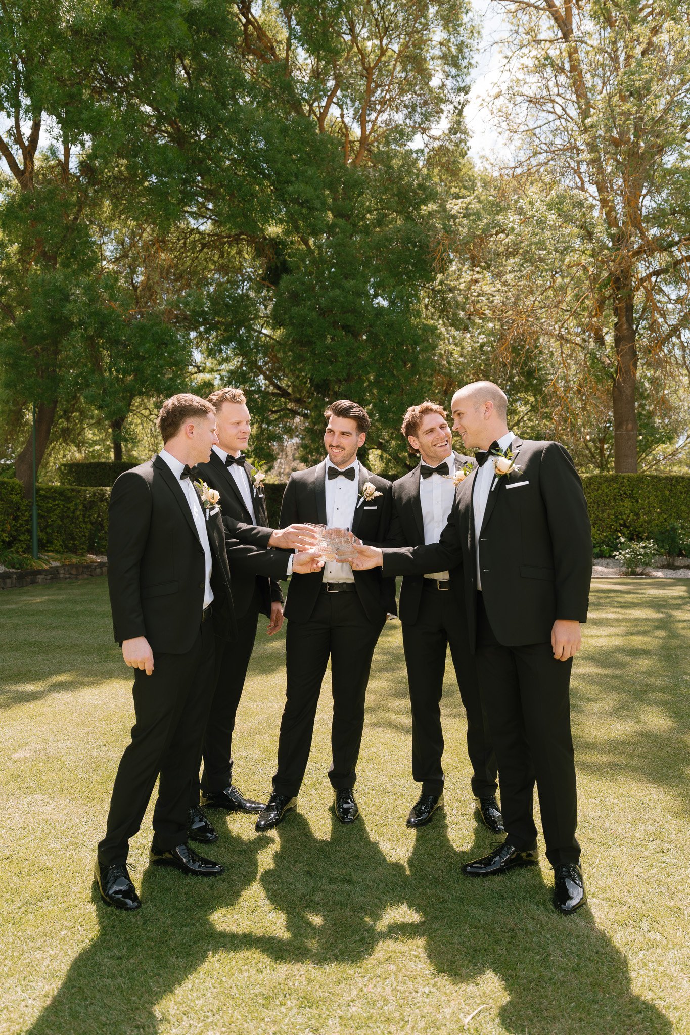 Group of six men in tuxedos celebrating outdoors, holding a glass award together, with trees and sunlight in the background.