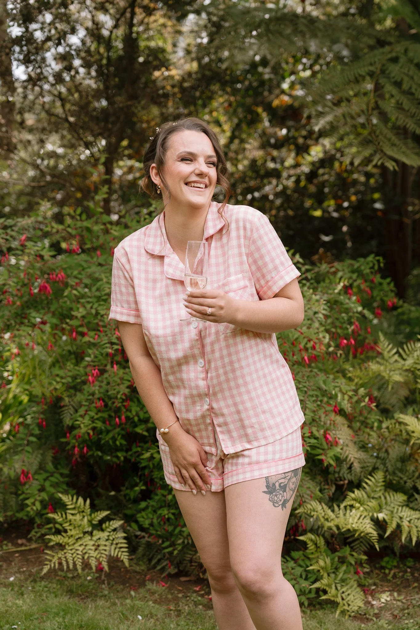 A woman in pink checkered pajamas holding a champagne glass, standing outdoors among greenery and flowering bushes, smiling cheerfully.