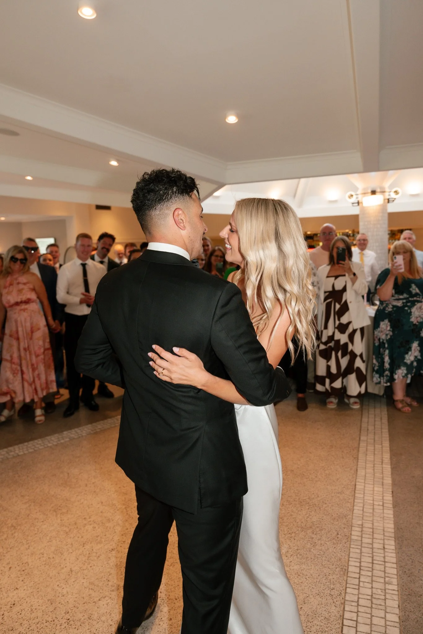 A couple dancing together at their wedding reception, surrounded by guests taking photos and watching them dance.
