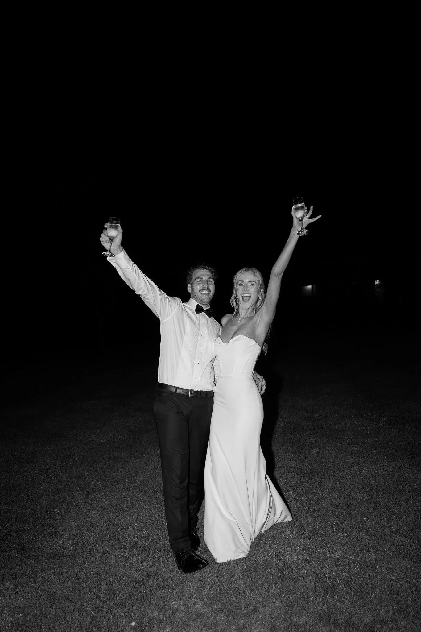 A joyful couple in wedding attire celebrating outdoors at night, holding glasses of champagne. The man wears a tuxedo with a bow tie, and the woman wears a strapless wedding dress with her arms raised in celebration.