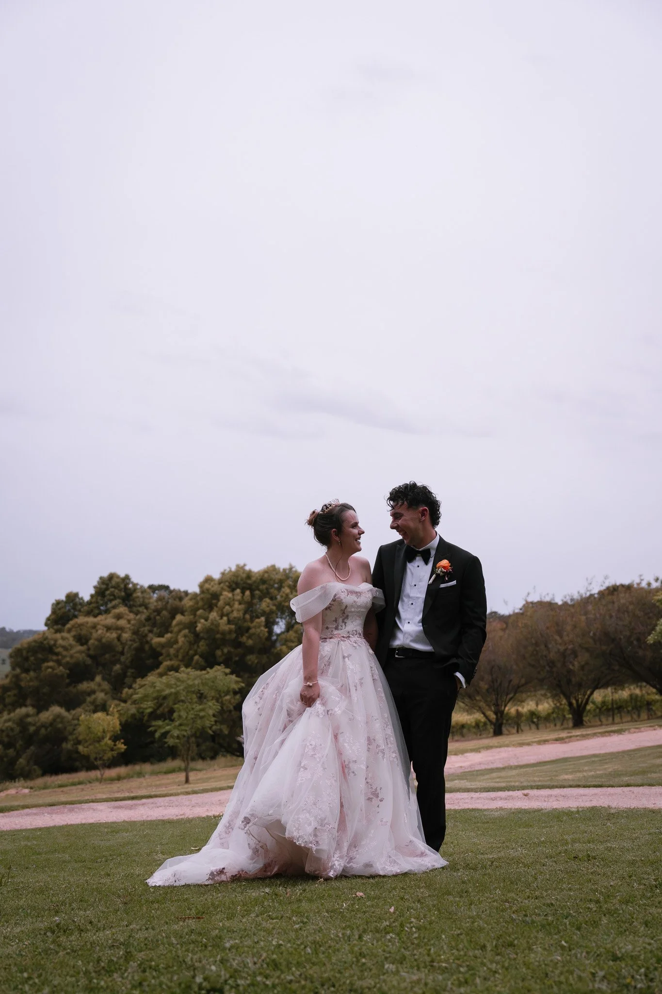 A bride and groom smiling at each other outdoors with trees and an overcast sky in the background.