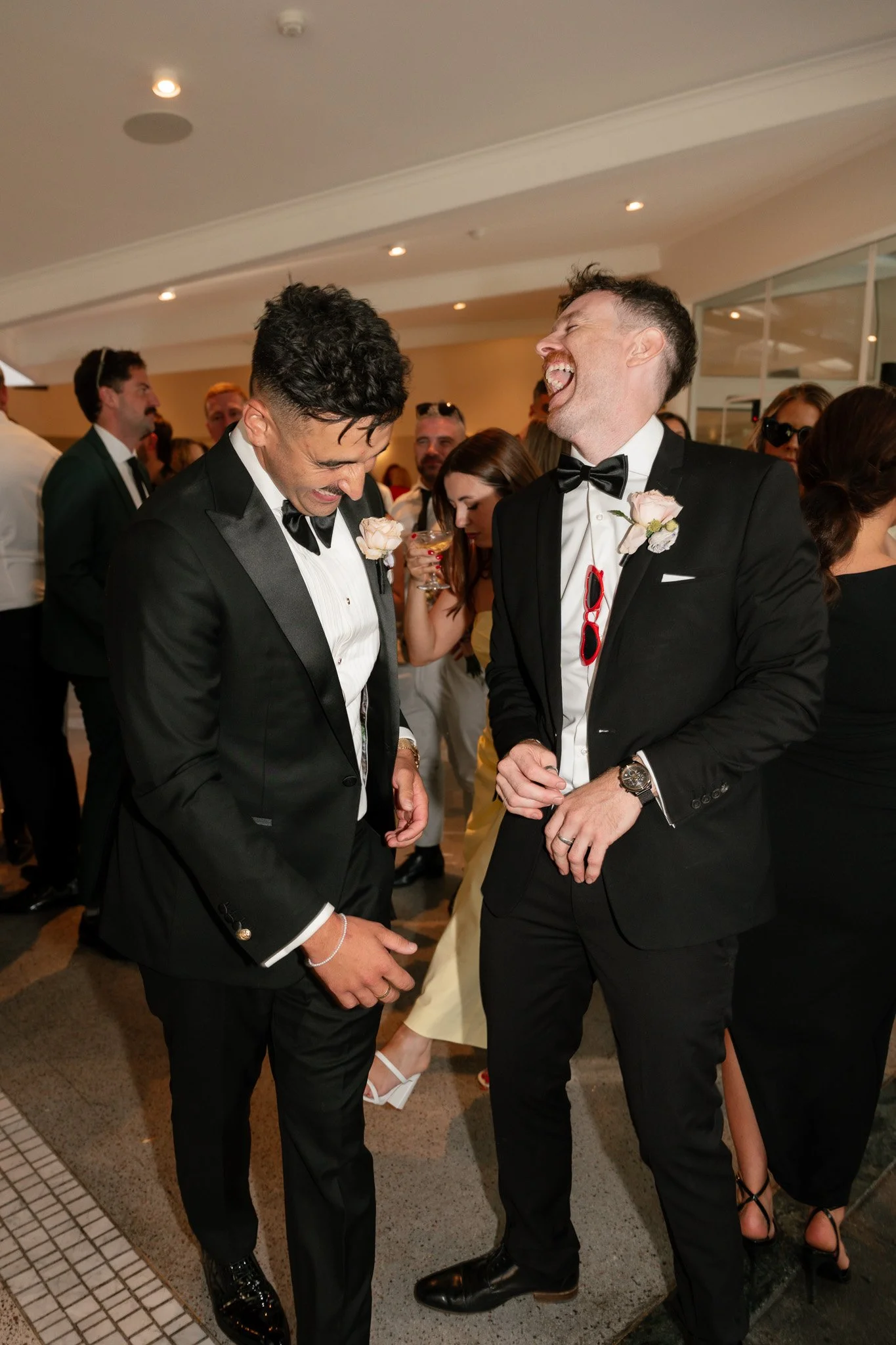 Two men dressed in tuxedos laughing and enjoying a moment at a formal event, surrounded by other guests.