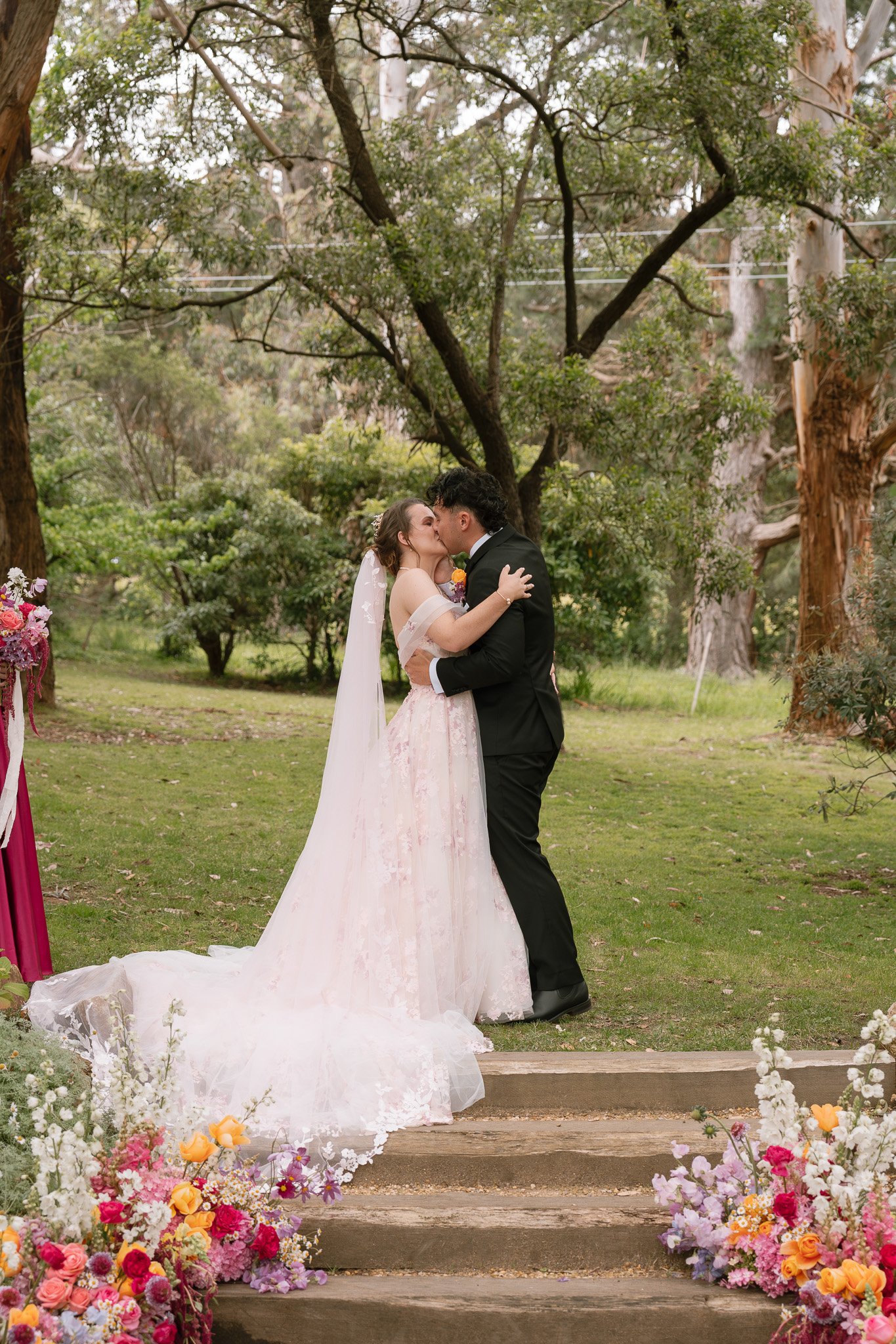A bride and groom kiss during their outdoor wedding ceremony, surrounded by trees and colorful flowers.