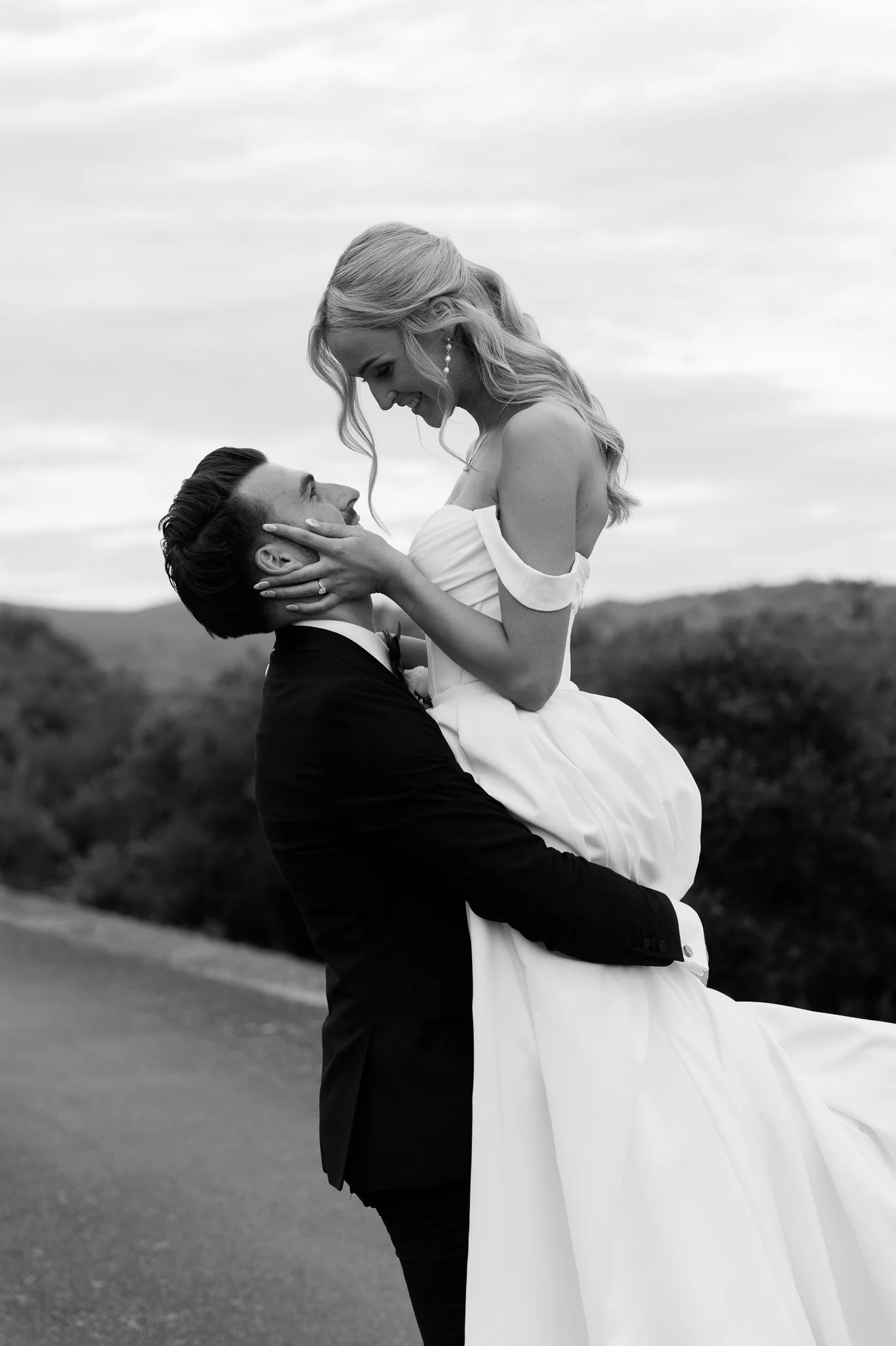 A black and white photo of a couple on their wedding day, with the man lifting the woman in his arms as they smile and look at each other outdoors.