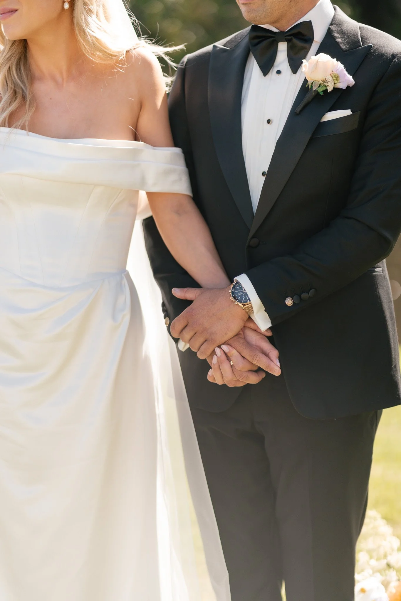 Close-up of a bride and groom holding hands during a wedding ceremony, with the bride in a white off-shoulder gown and the groom in a black tuxedo with a bow tie, outdoors.
