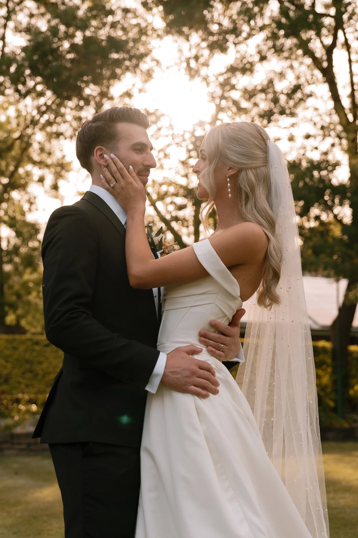 A bride and groom are embracing outdoors during sunset, with the bride gently touching the groom's face, both gazing into each other's eyes, surrounded by trees.