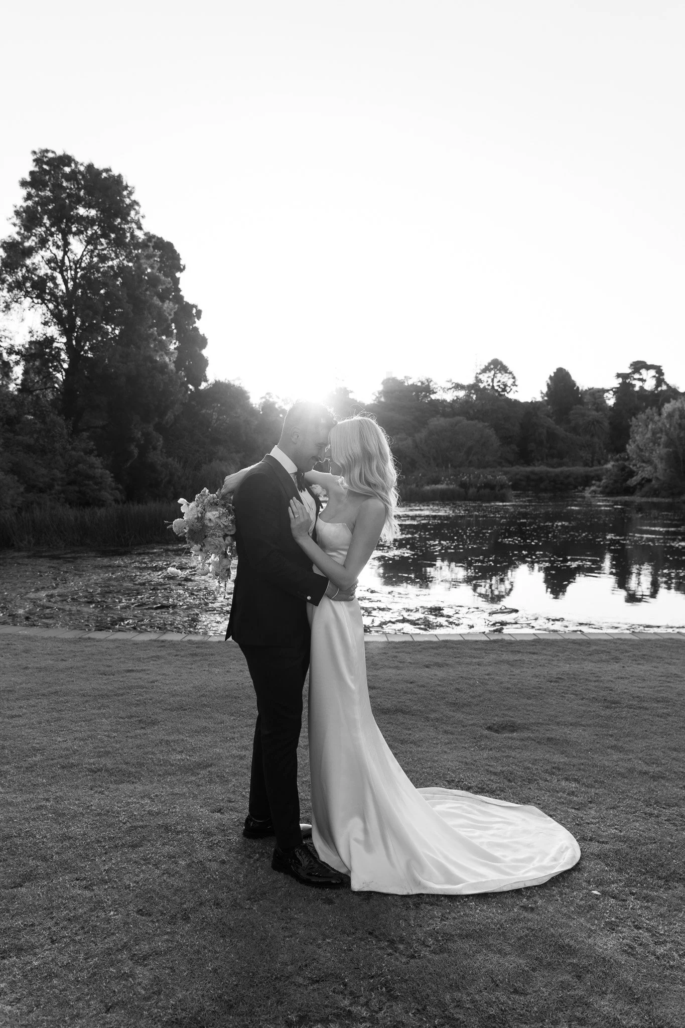 A black and white photo of a bride and groom standing close together outdoors near a body of water, at sunset, with trees in the background. The bride is holding a bouquet and wearing a long, elegant wedding dress, while the groom is in a suit. They 