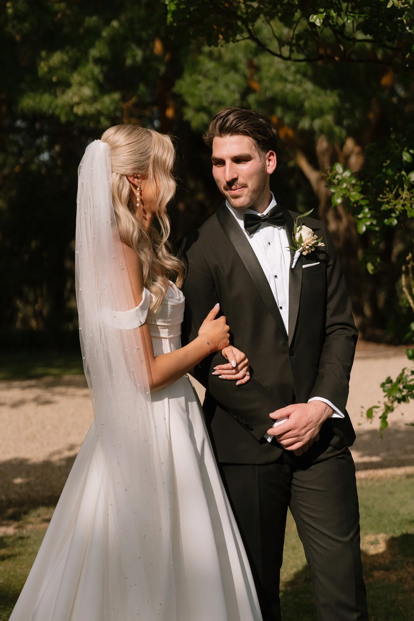 A bride and groom standing outdoors, holding hands, with trees in the background. The bride wears a white wedding gown and veil; the groom wears a black tuxedo with a bowtie.