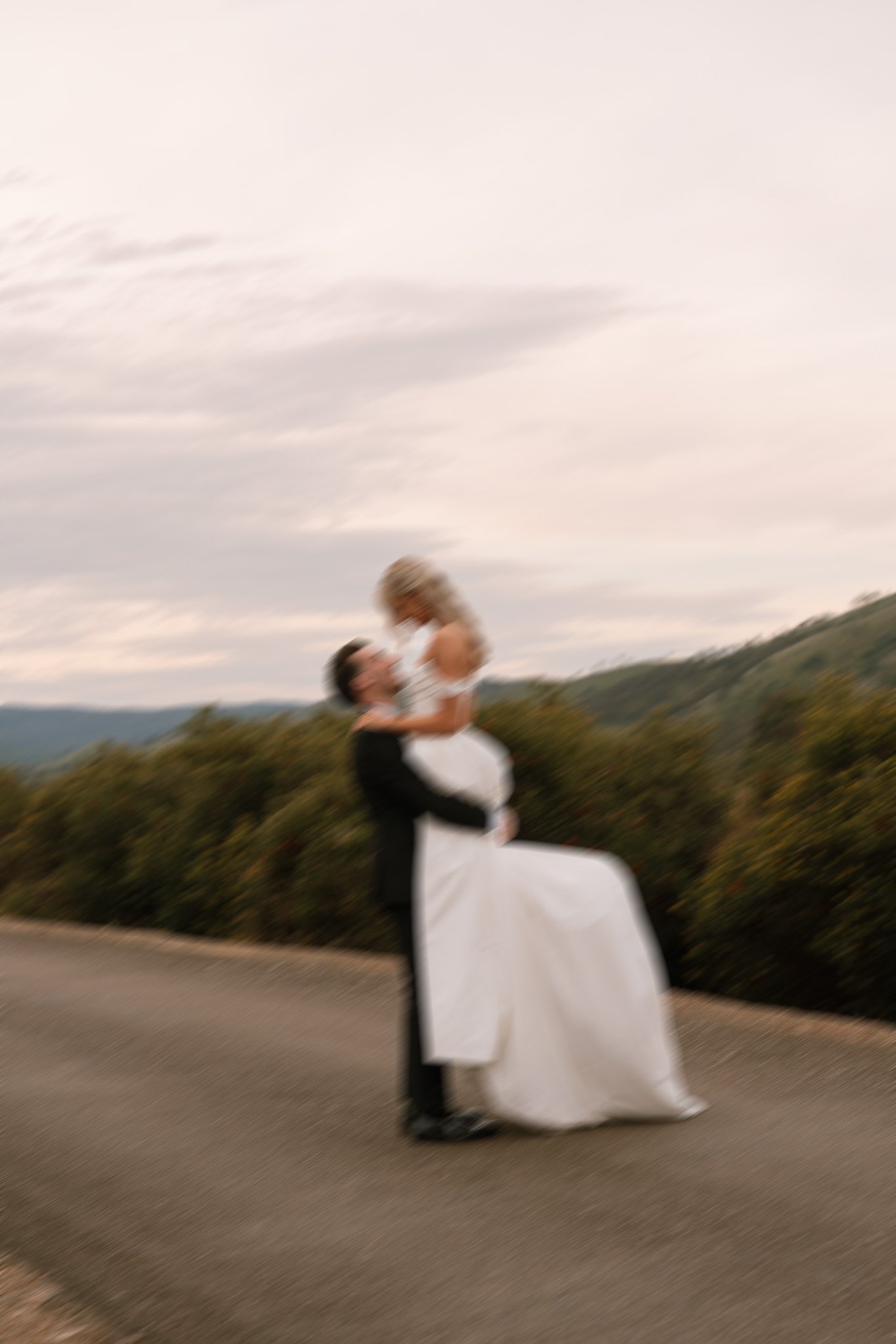 A blurry photo of a couple in wedding attire, with the groom holding the bride in his arms outdoors on a road, with hills and a cloudy sky in the background.