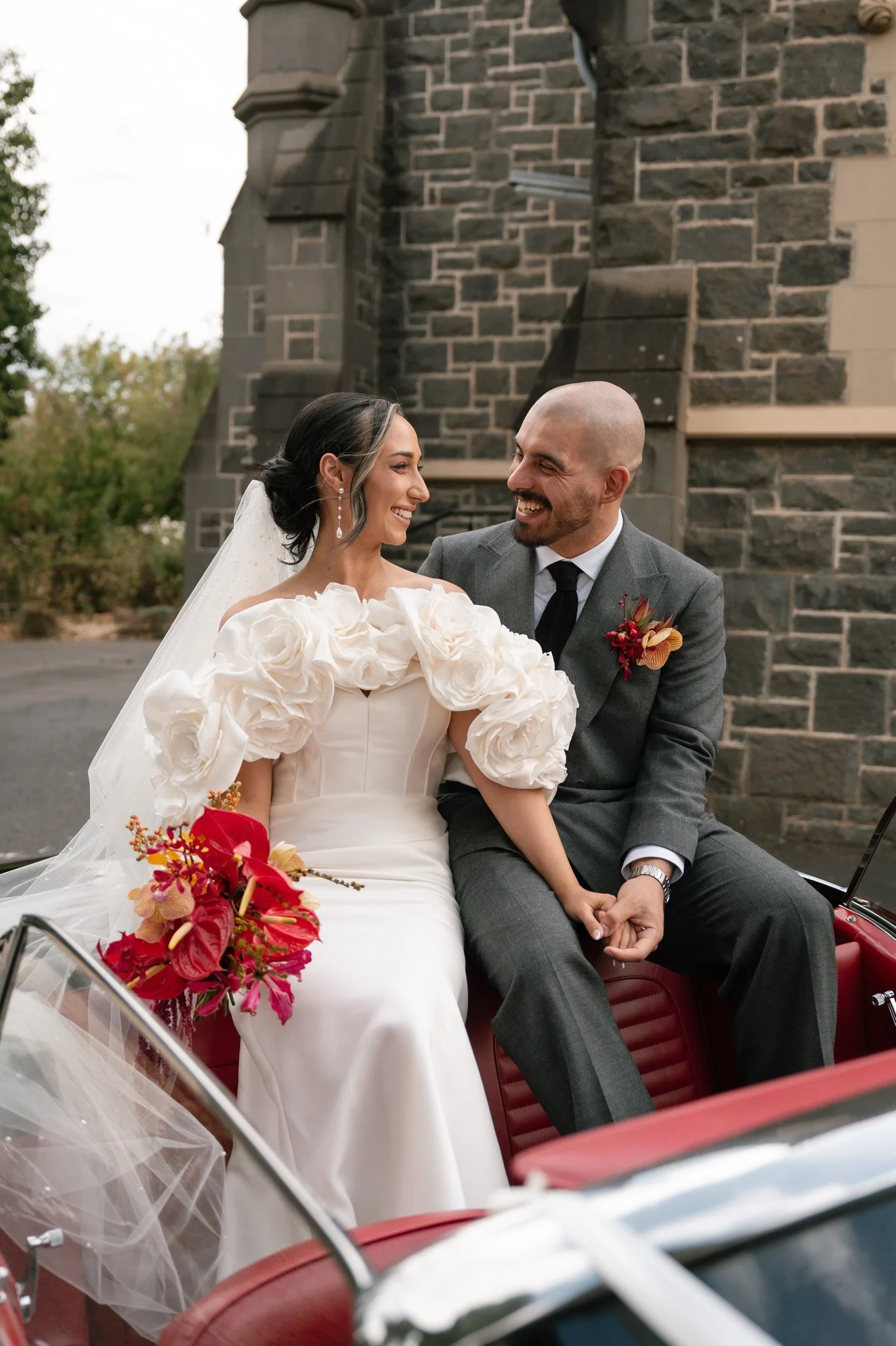 Bride and groom sitting together in a vintage car, smiling and holding hands, with a stone building in the background.