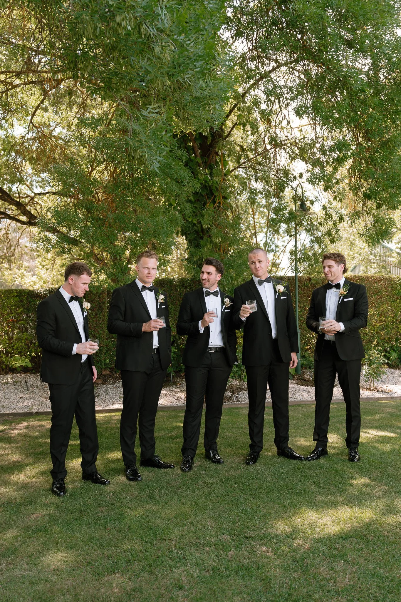 Group of five men in tuxedos with bow ties and white shirts, holding glasses, standing outdoors on grass near a large leafy tree during daytime, possibly at a formal event or wedding.