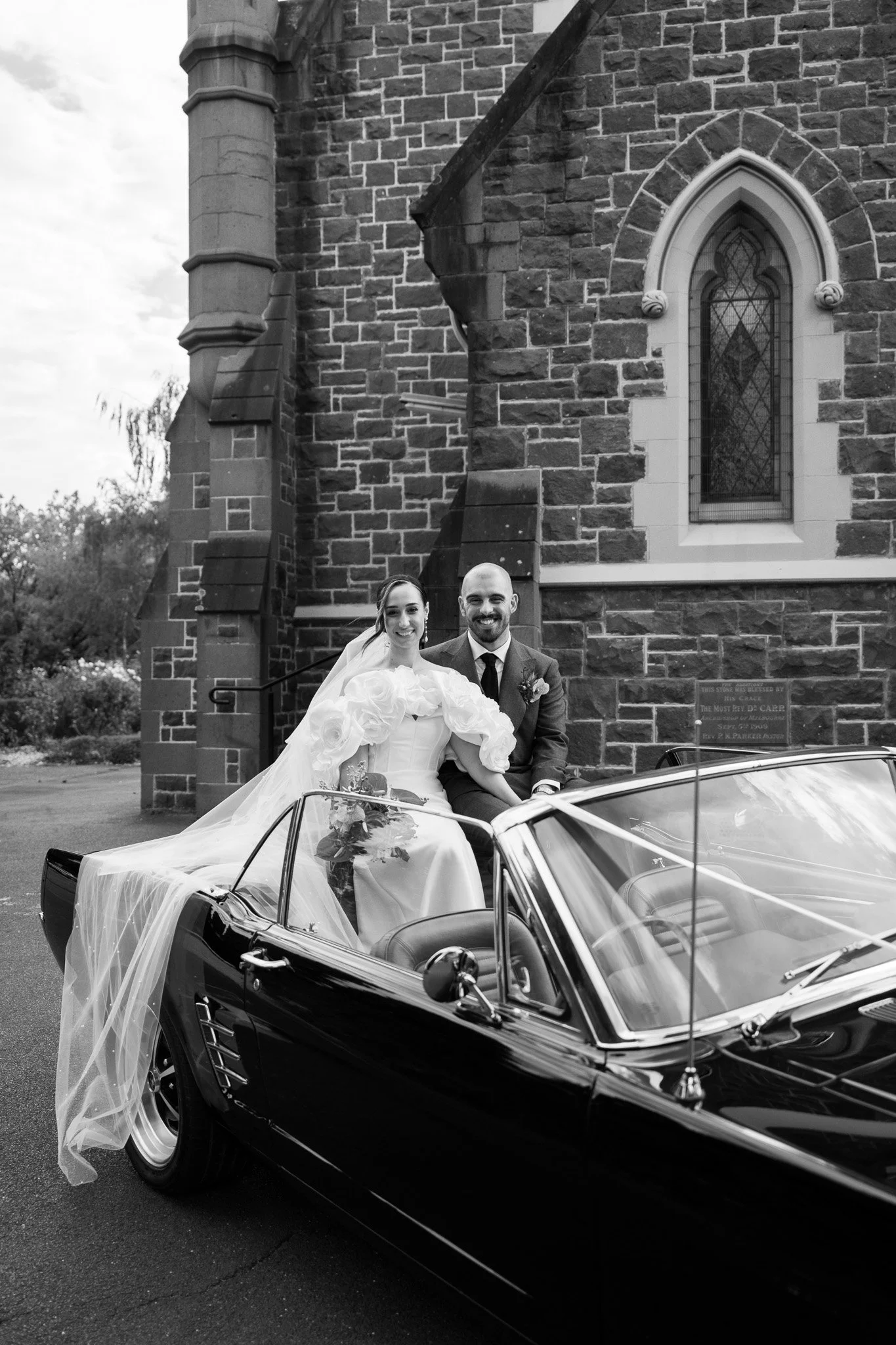 A bride and groom sitting in a convertible car, smiling, outside a stone church on their wedding day, in black and white.