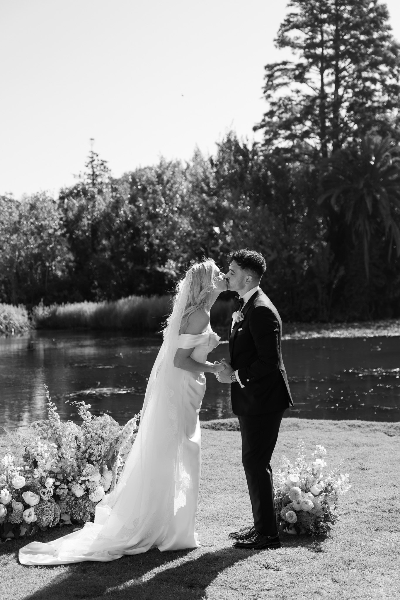 Black and white photo of a bride and groom sharing a kiss outdoors by a pond, holding hands, with flowers at their feet and trees in the background.