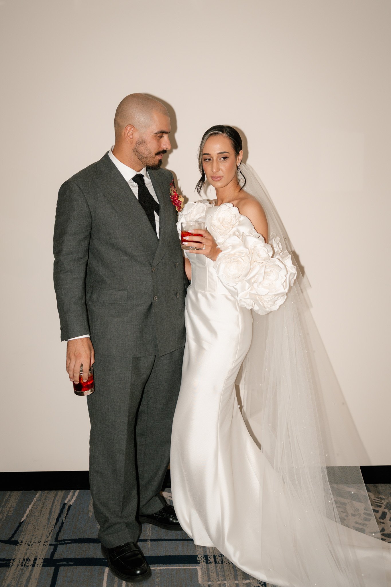 A bride and groom standing together, each holding a glass with red beverage, posing against a plain wall. The bride is wearing a white satin wedding gown with large floral embellishments on the sleeves, and a veil. The groom is dressed in a dark grey