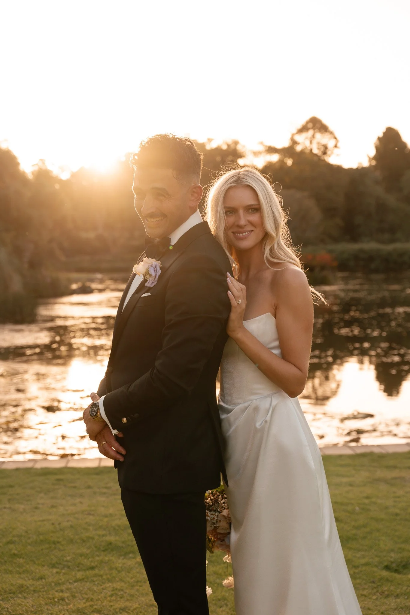 A newlywed couple stands by a river at sunset, with trees in the background. The groom wears a black tuxedo with a white shirt and black bow tie, and the bride wears a strapless white wedding gown. The sunlight creates a warm glow around them.