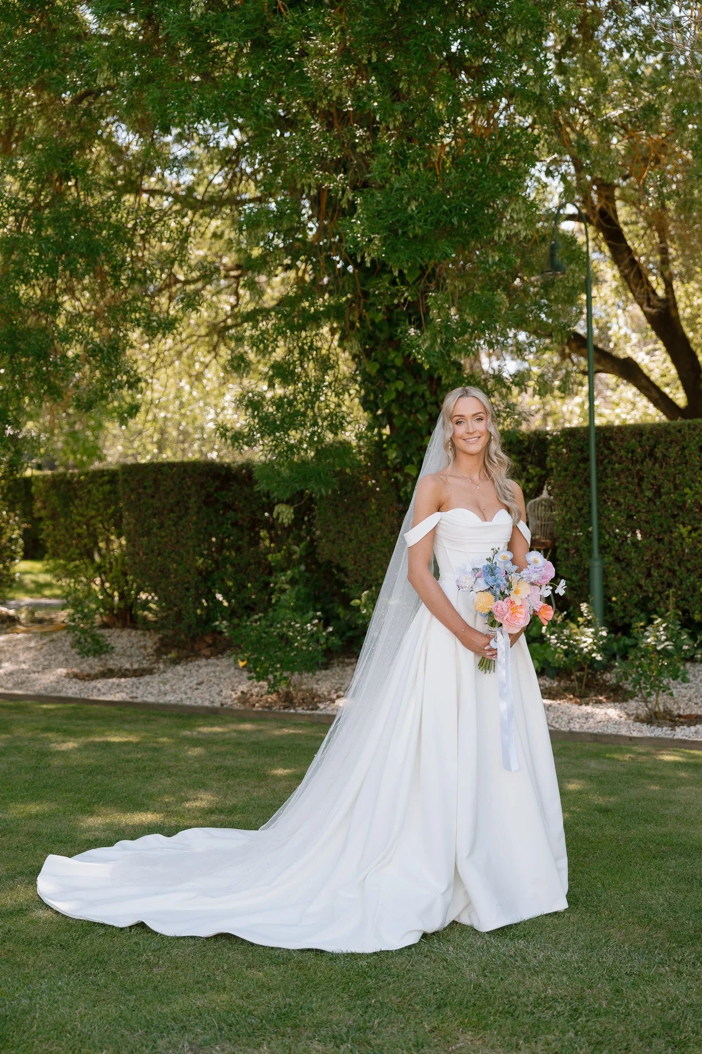 Bride in a white wedding dress holding a colorful bouquet, standing outdoors on a lawn with trees and bushes in the background.
