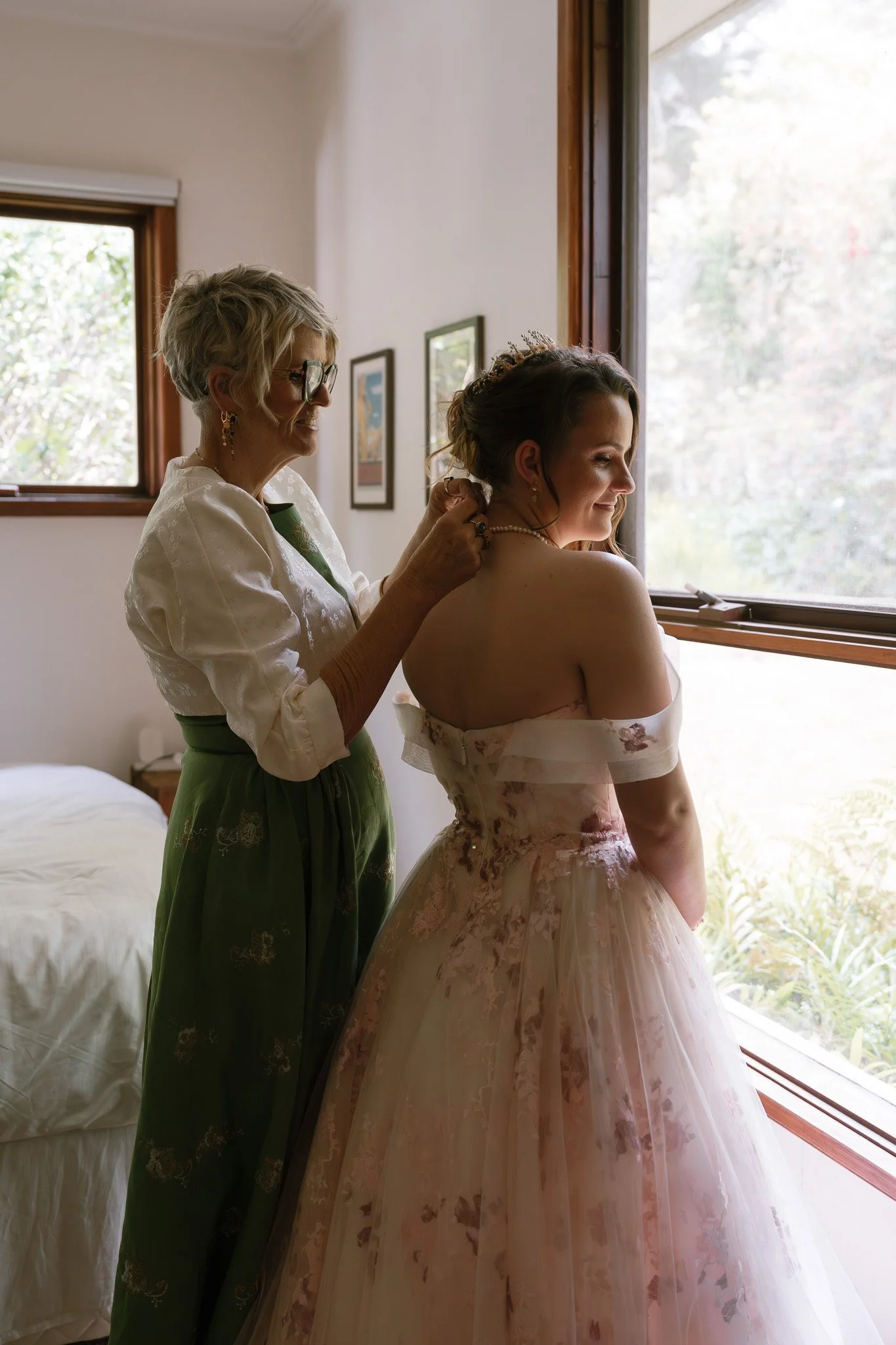 A woman helping another woman with her necklace by a window in a room, with framed pictures on the wall, a bed, and trees outside the window.