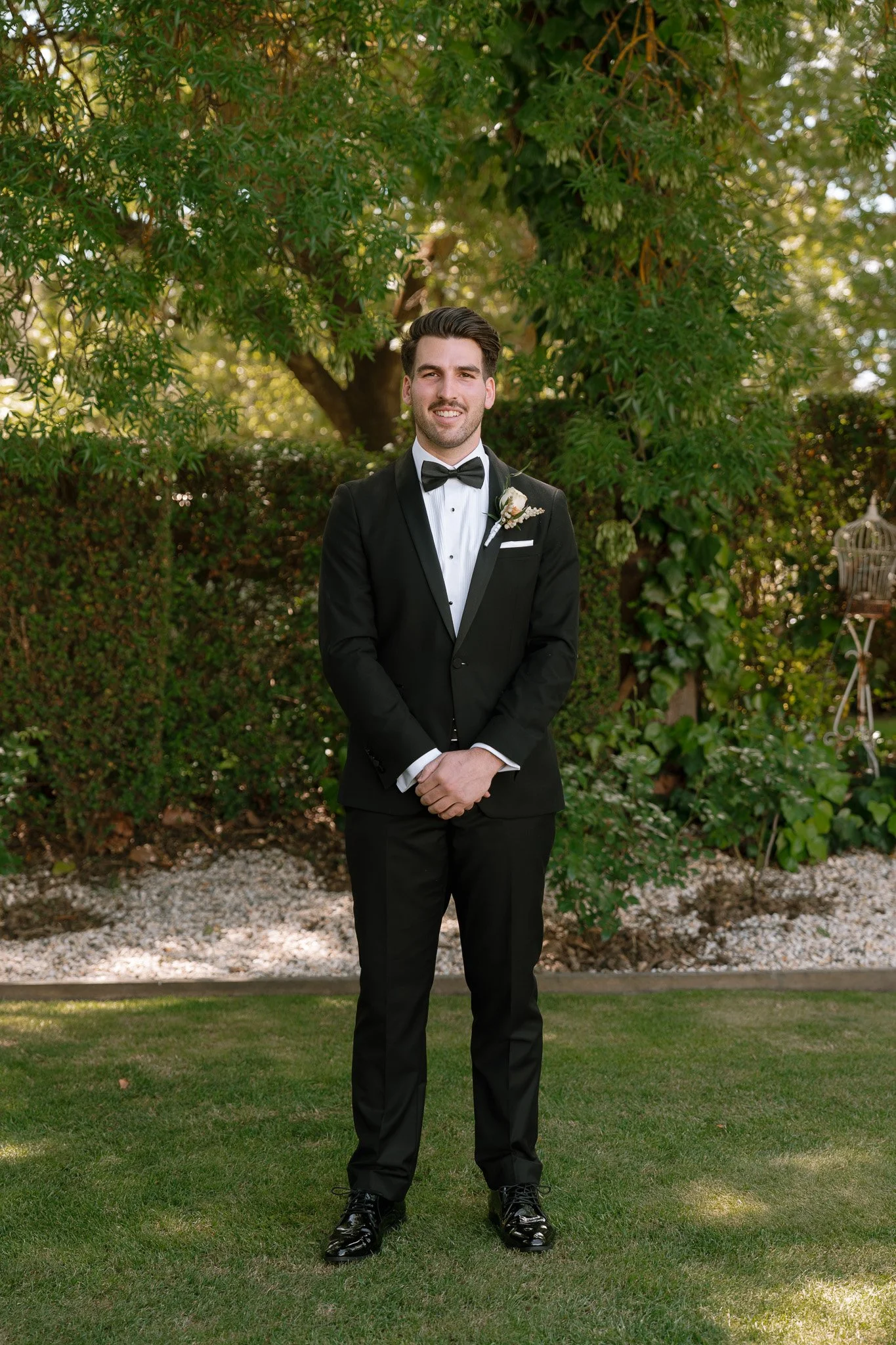 A groom in a black tuxedo with a bow tie standing outdoors on a grassy area in front of green trees and bushes.