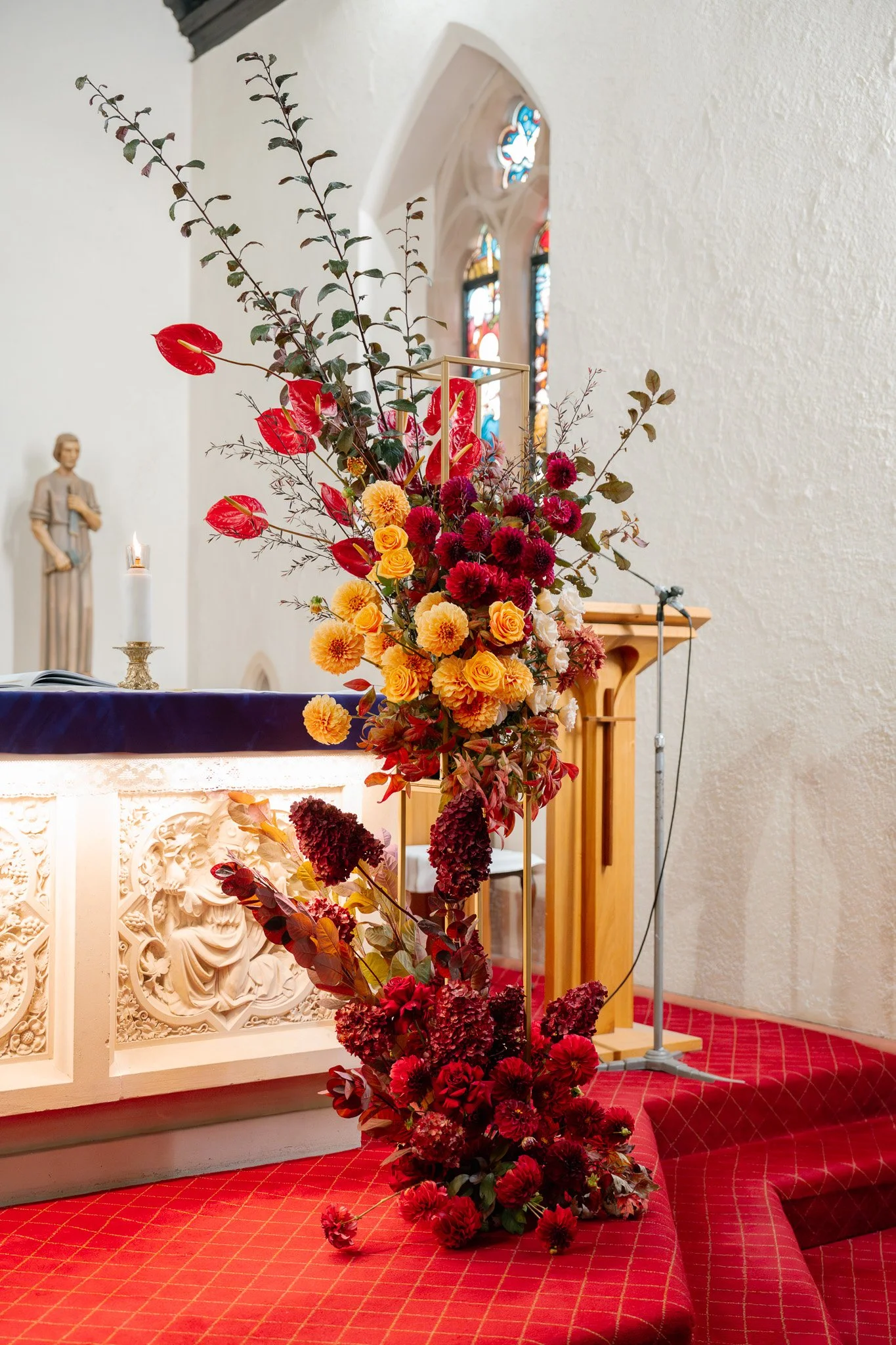 A large floral arrangement with red, yellow, and dark purple flowers and green foliage on a table in a church, with stained glass windows and a statue in the background.